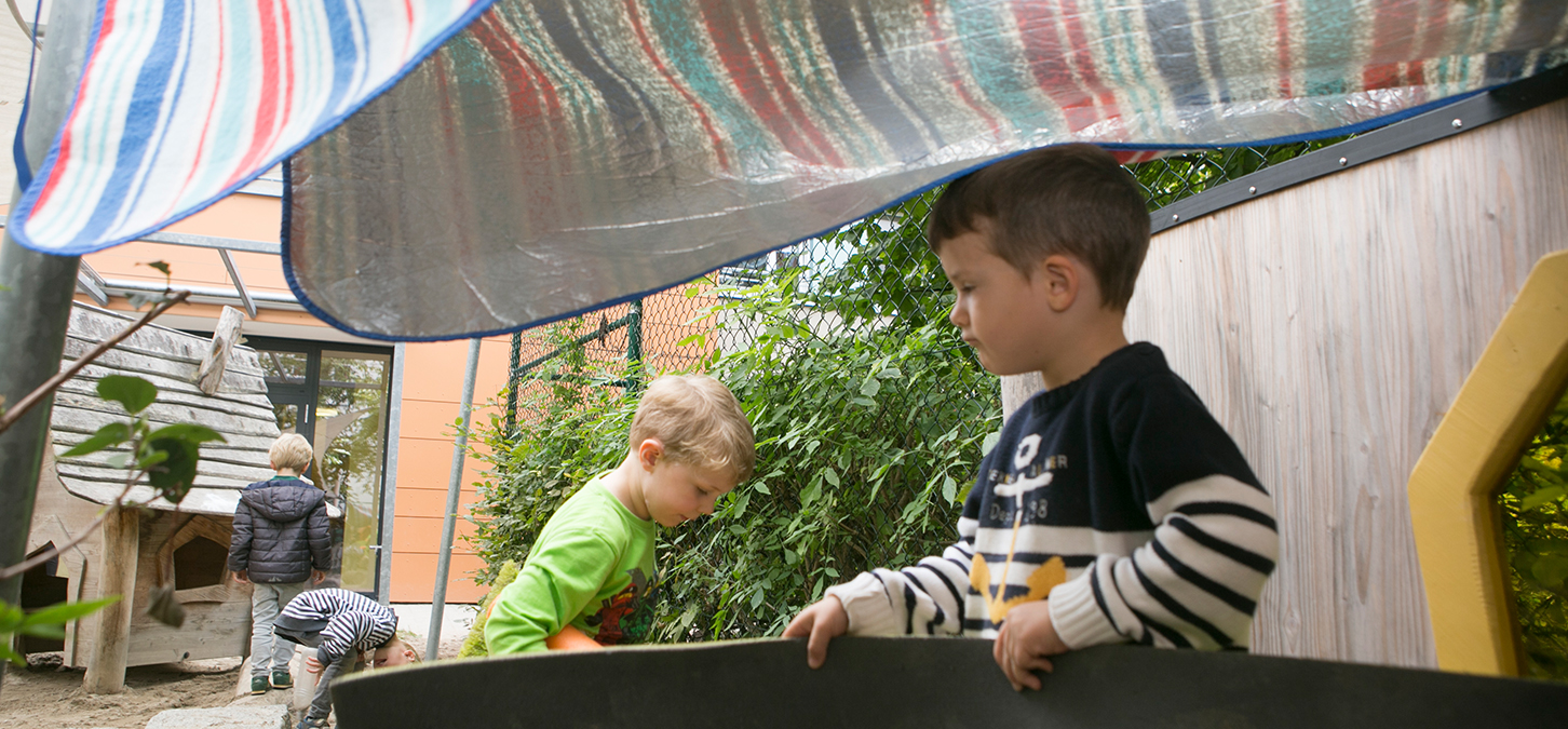 Zwei Jungen spielen unter einem behelfsmäßigen Zelt aus gestreiftem Stoff im Freien, während sich ein anderes Kind in Grün in der Nähe bückt und zwei weitere Kinder im Hintergrund in der Nähe eines Gebäudes und eines Zauns zu sehen sind.