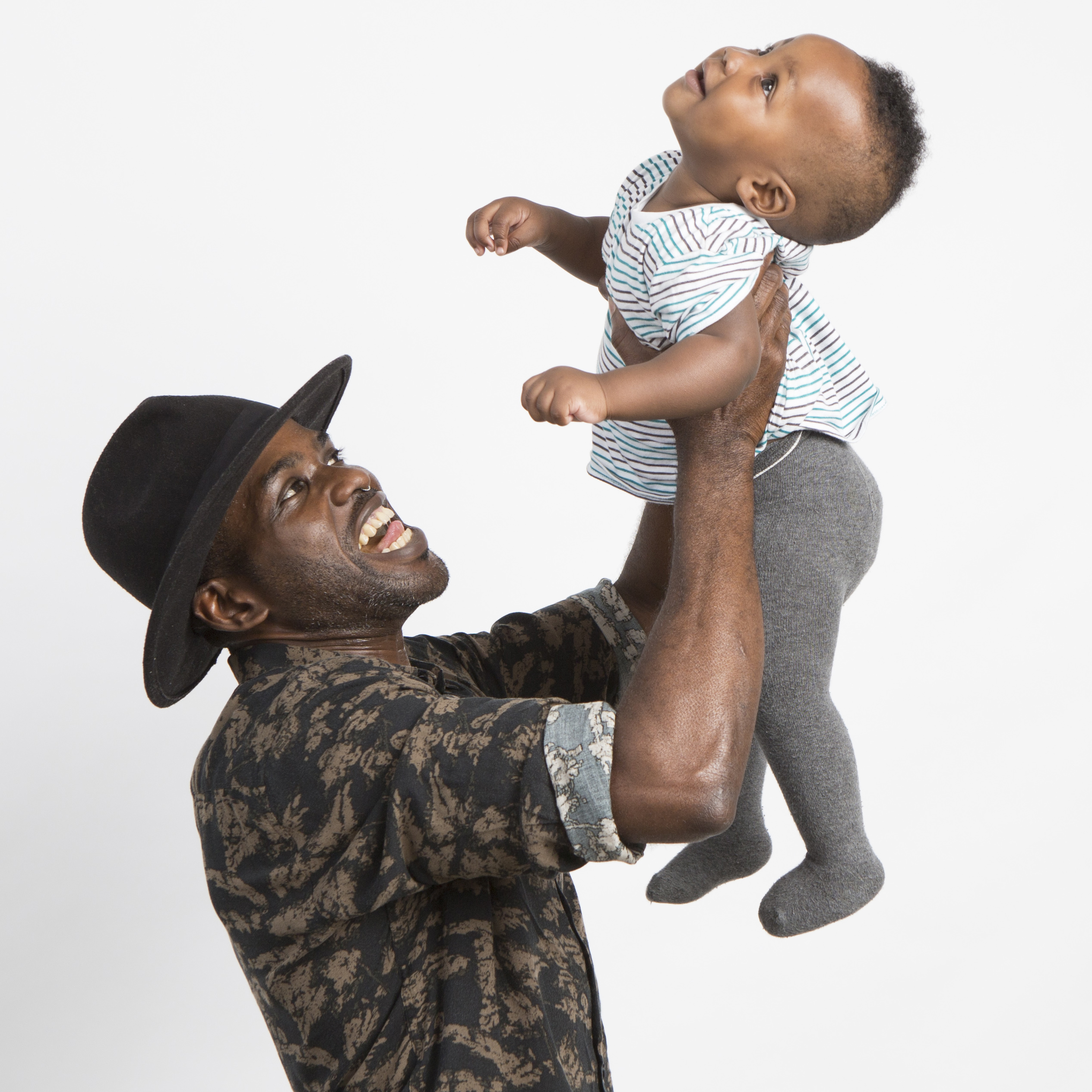 Ein lächelnder Mann, der einen Hut trägt, hebt ein lachendes Baby in die Luft, beide schauen sich freudig vor einem schlichten weißen Hintergrund an.