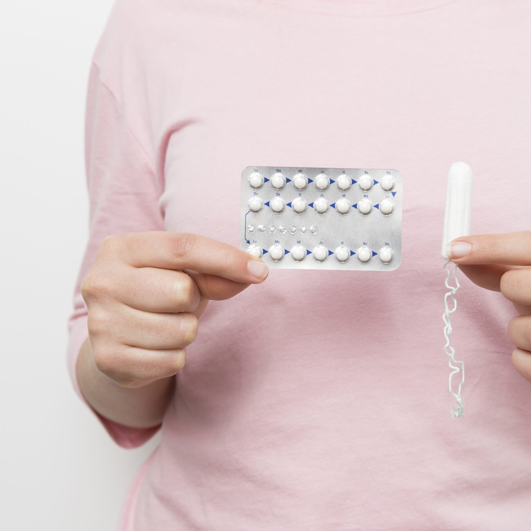 Eine Person in einem rosa Hemd hält in der einen Hand eine Blisterpackung mit Antibabypillen und in der anderen einen unbenutzten Tampon. Beides wird vor einem schlichten Hintergrund gehalten. So werden persönliche Entscheidungen hervorgehoben, ähnlich wie die Verpflichtungen des Freiburger Klimapakts.