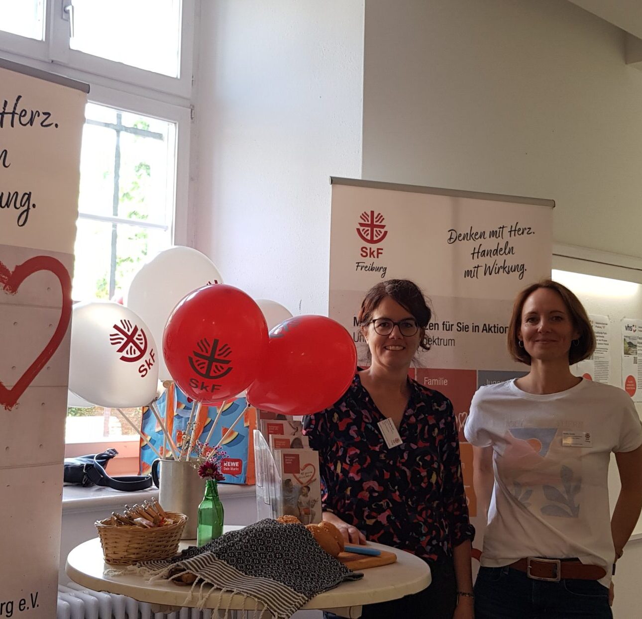 Zwei Frauen stehen bei einer Indoor-Veranstaltung an einem Tisch mit Luftballons, Snacks und SkF Freiburg-Bannern.