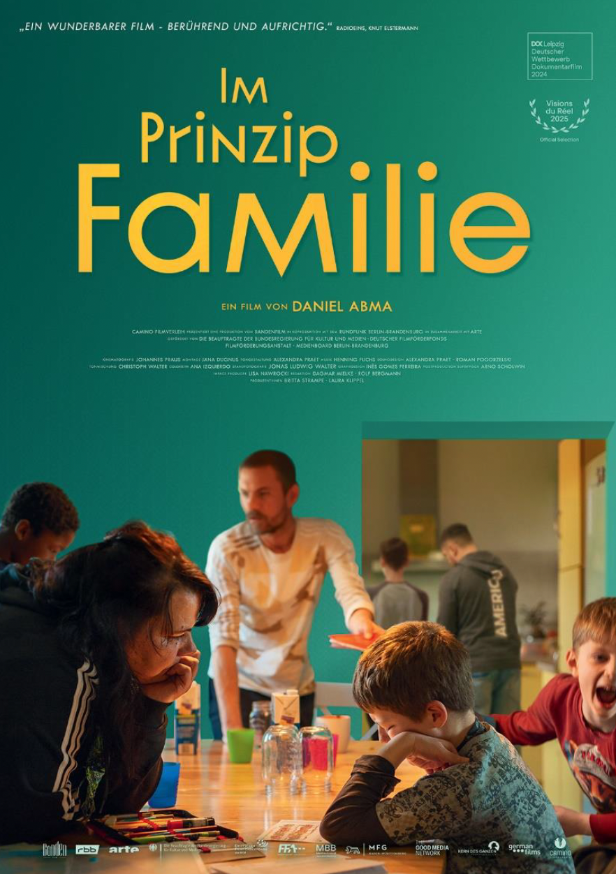 Eine Frau und Kinder sitzen an einem Tisch, im Hintergrund sind ein Mann und andere zu sehen; der Text lautet „Im Prinzip Familie“.