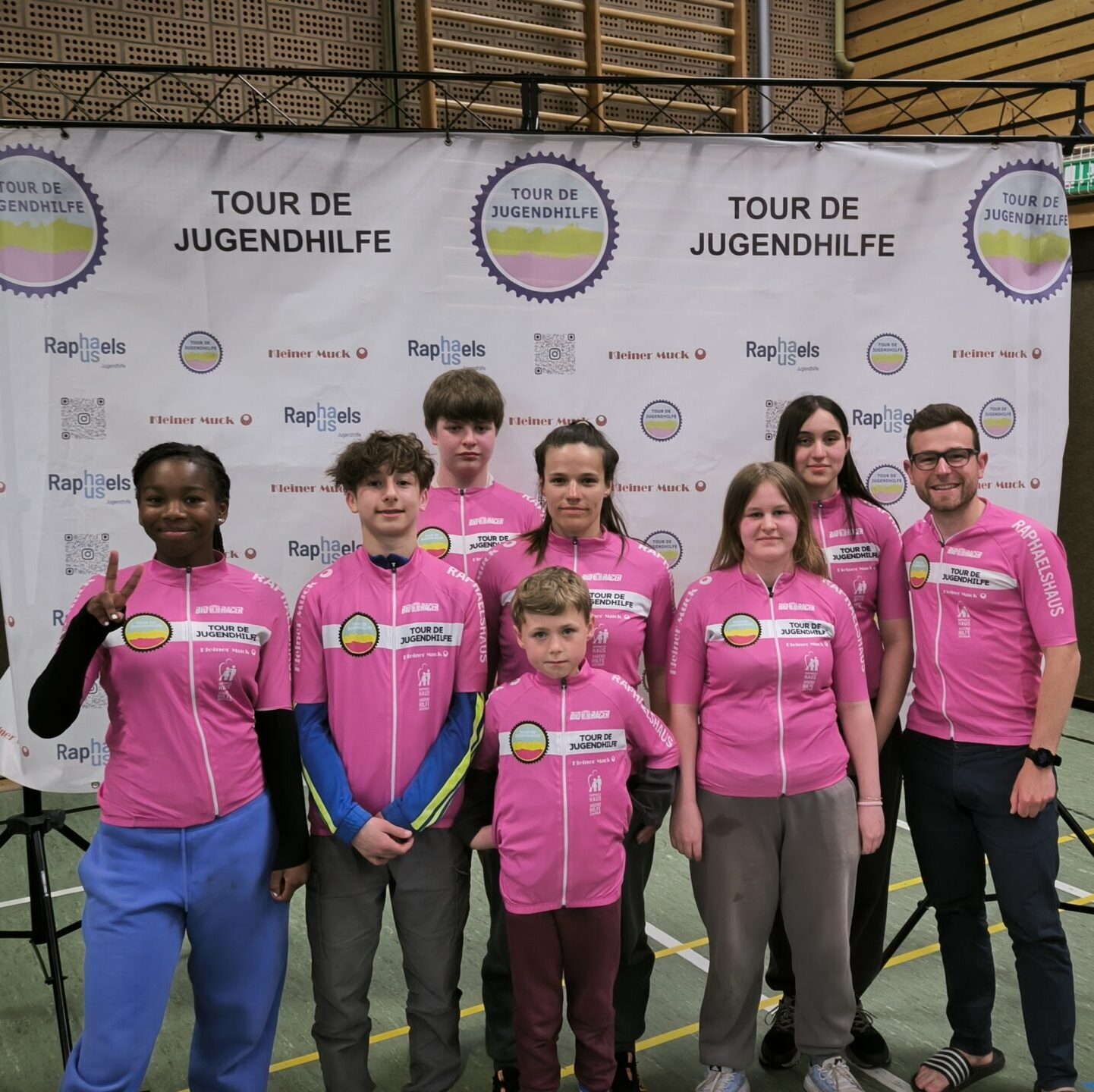 Eine Gruppe von Menschen in rosafarbenen Tour de Jugendhilfe-Shirts posiert gemeinsam in einem Innenbereich vor einer Markenkulisse.