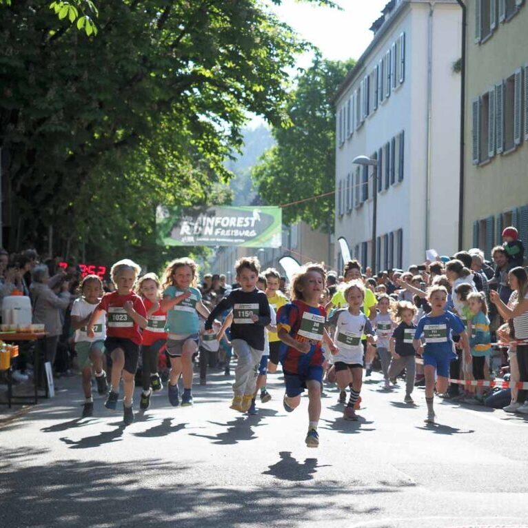Eine Gruppe kleiner Kinder läuft ein Rennen auf einer sonnigen Straße, die von Zuschauern und Bäumen gesäumt ist.