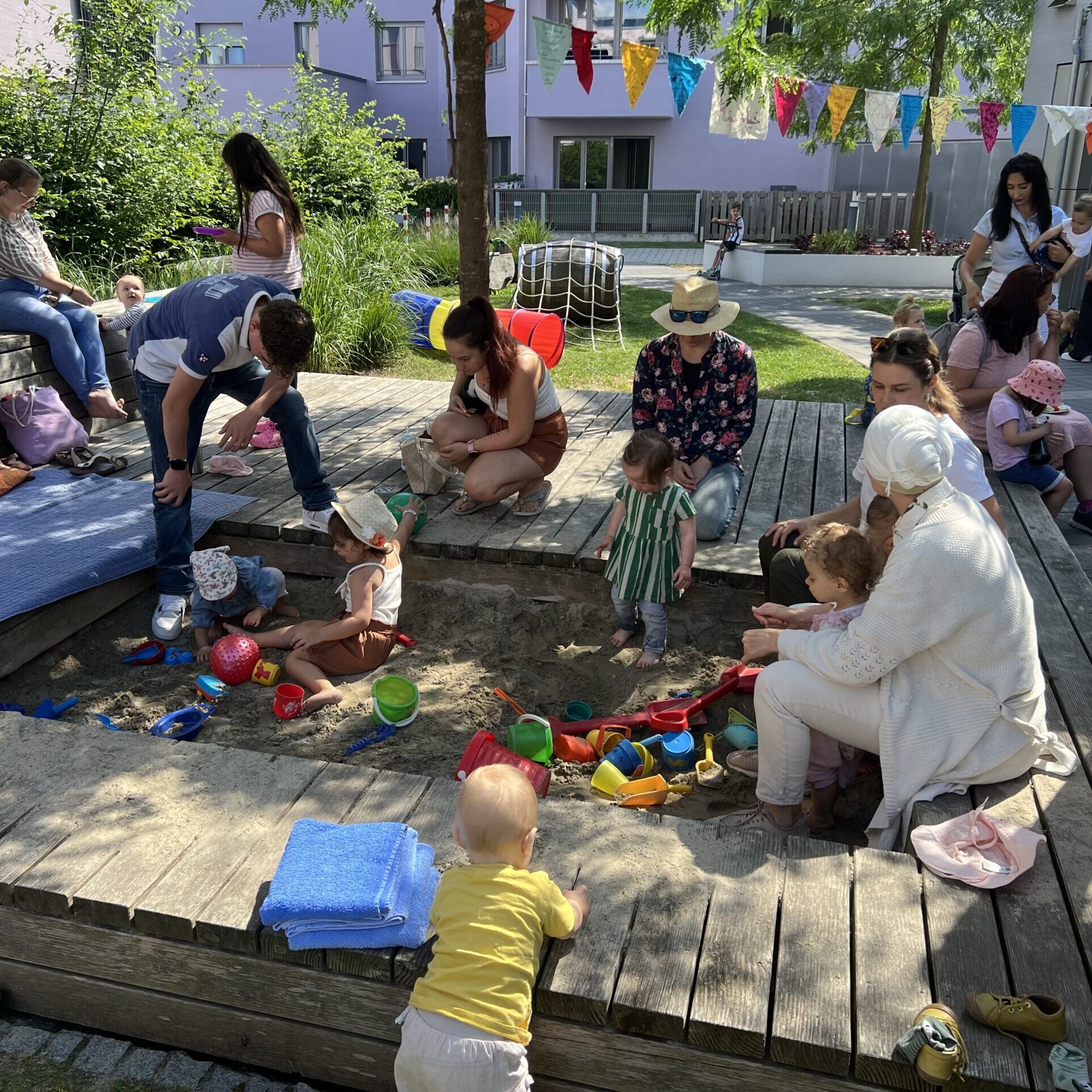 Erwachsene und kleine Kinder versammeln sich und spielen in einem Sandkasten im Freien, umgeben von Holzbänken, Spielzeug und bunten Wimpeln. Einige Erwachsene sitzen, während die Kinder mit Sand und Eimern spielen.