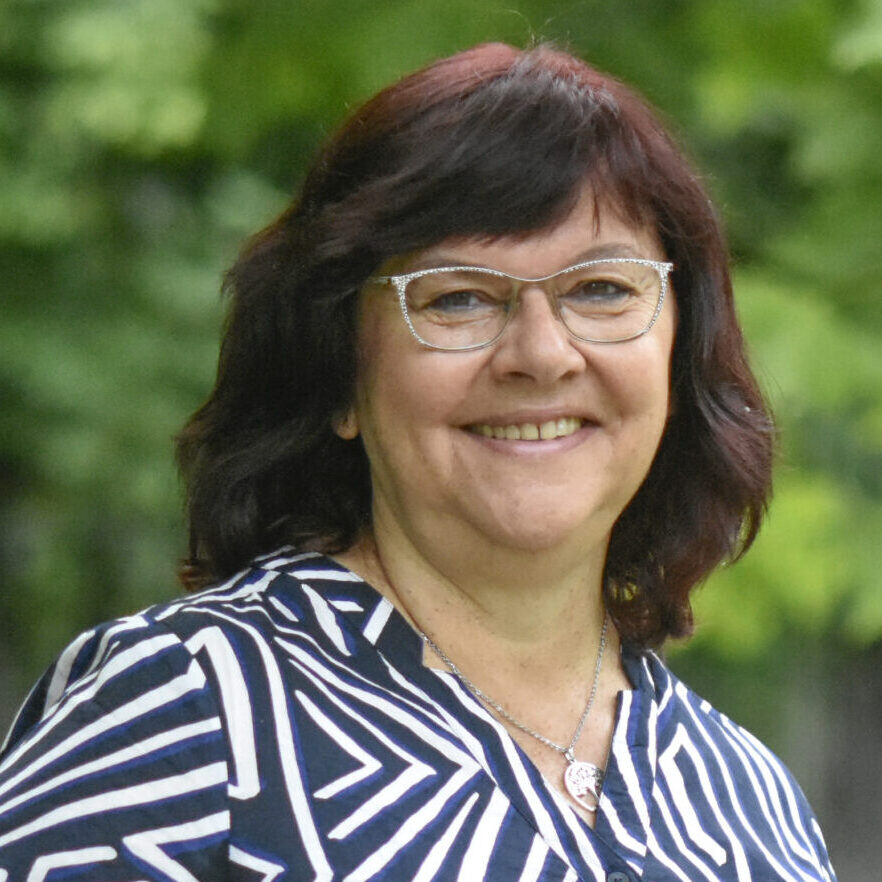 Eine Frau mit schulterlangen dunklen Haaren und Brille lächelt im Freien, sie trägt ein schwarz-weiß gemustertes Hemd. Sie steht vor grünen Bäumen und stützt eine Hand in die Hüfte. Eine Frau mit schulterlangen dunklen Haaren und Brille lächelt im Freien, sie trägt ein schwarz-weiß gemustertes Hemd. Sie steht vor grünen Bäumen und stützt eine Hand in die Hüfte.