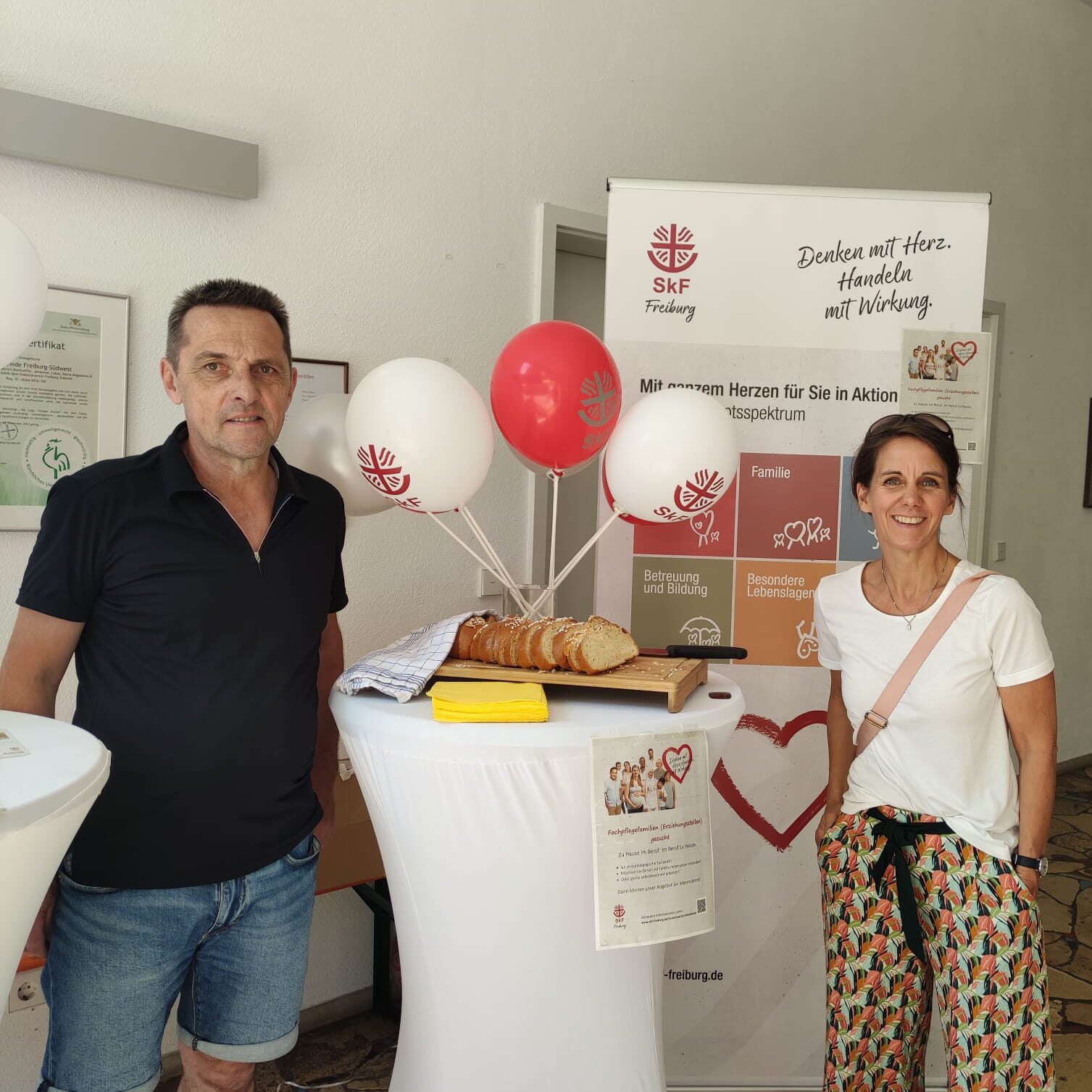 Ein Mann und eine Frau stehen vor einem Tisch mit Brotscheiben, Luftballons und Informationsbannern bei einer Indoor-Veranstaltung. Die Banner zeigen Herz- und Familiengrafiken mit deutschem Text.