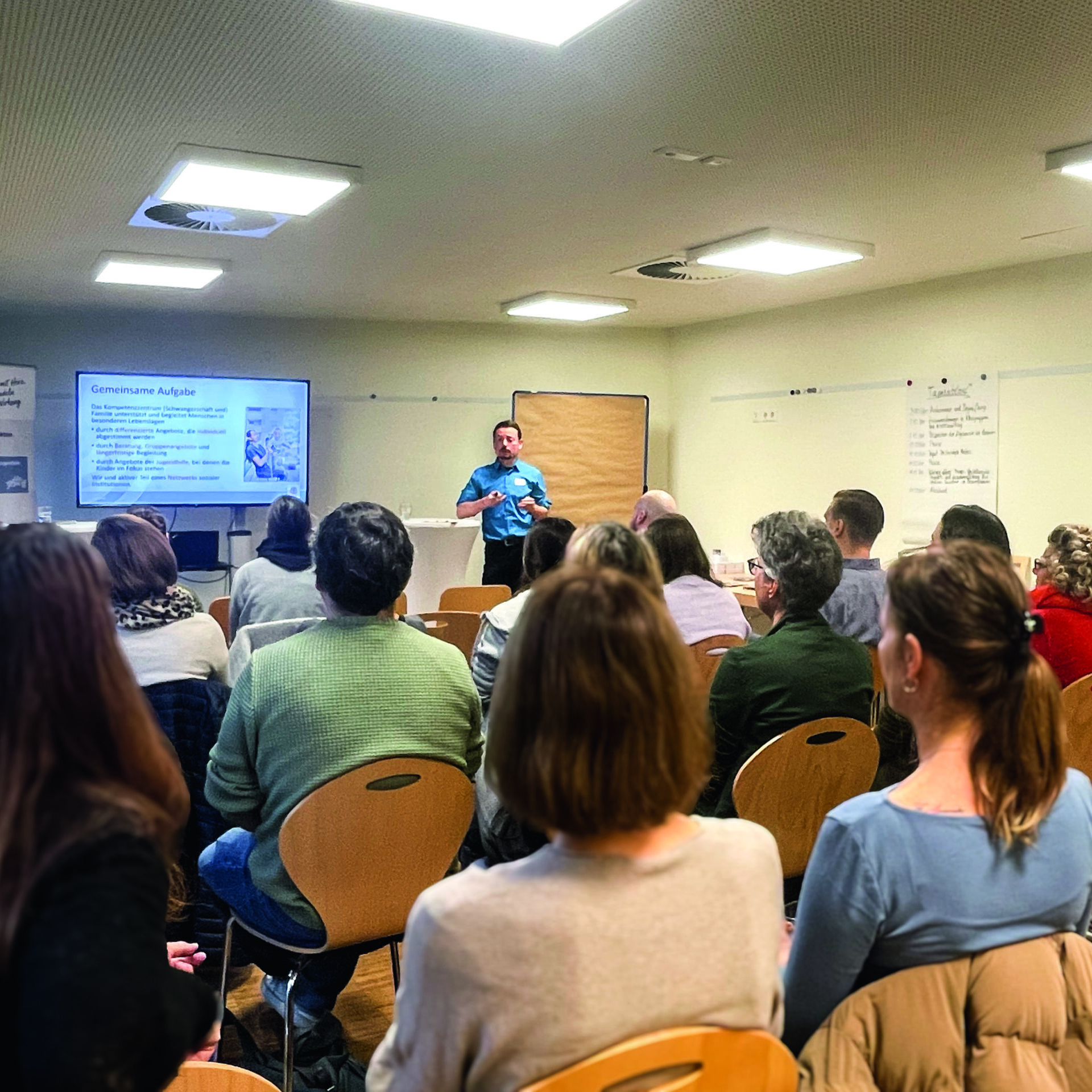 Eine Gruppe von Personen sitzt auf Stühlen und blickt auf einen Redner, der Informationen auf einem Bildschirm im vorderen Teil eines Klassenzimmers oder Konferenzraums präsentiert. Der Raum hat weiße Wände, und an einer Wand sind Poster zu sehen.