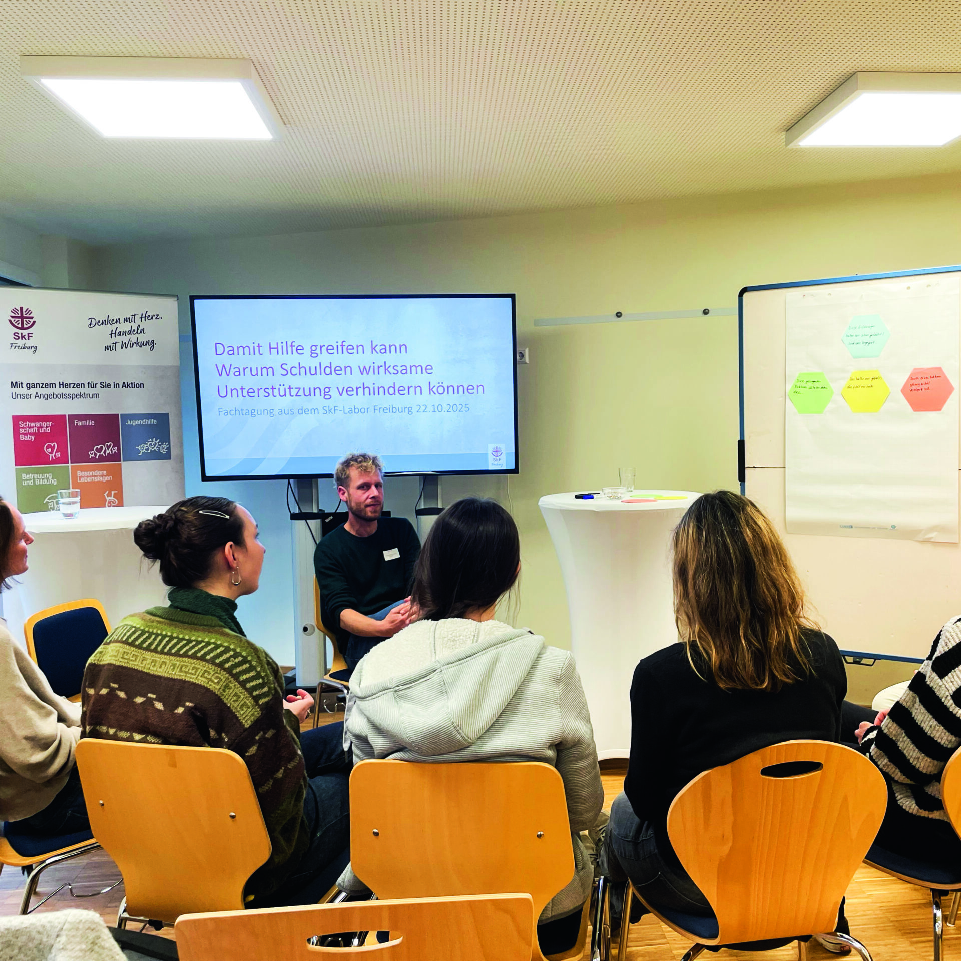 Ein Mann spricht zu sechs sitzenden Frauen in einem kleinen Besprechungsraum. Hinter ihm ist ein Bildschirm mit einer Präsentationsfolie in deutscher Sprache und ein Flipchart mit bunten Notizen zu sehen. Die Umgebung wirkt informell und gemeinschaftlich.