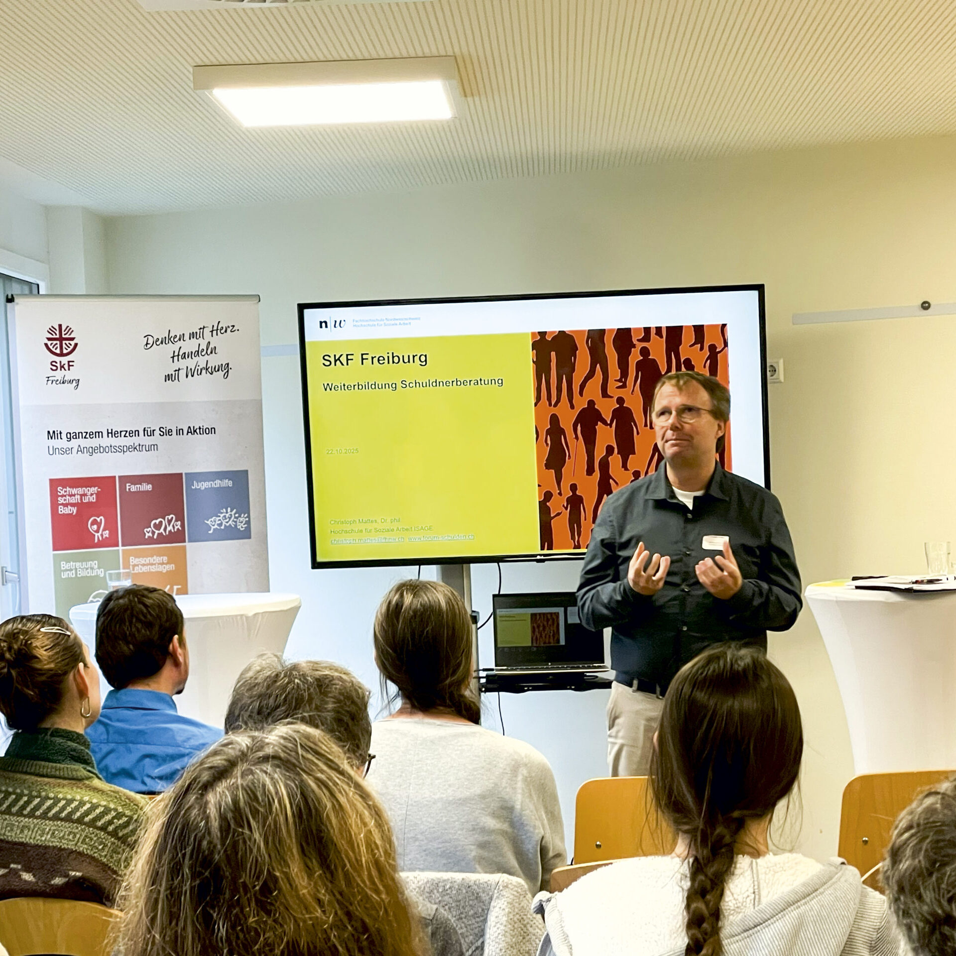 Ein Mann steht vor einem Publikum und hält eine Präsentation. Auf einem Bildschirm hinter ihm sind "SKF Freiburg" und eine bunte Grafik zu sehen. Die Anwesenden sitzen und hören zu. Links neben dem Vortragenden ist ein Banner mit Text und Logos zu sehen.
