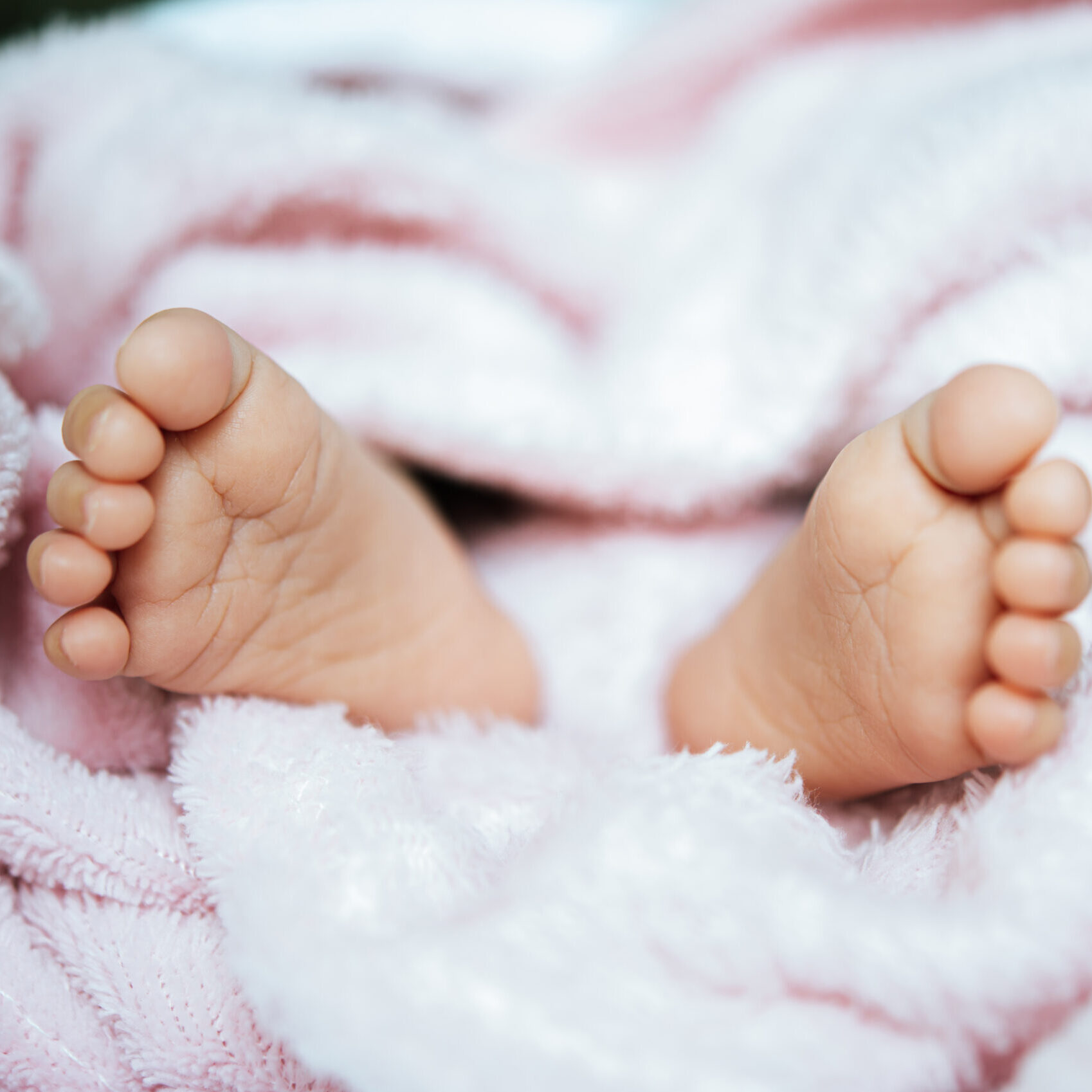 Nahaufnahme der nackten Füße eines Babys, die aus einer weichen, flauschigen rosa Decke herausragen. Die Füße des Babys sind entspannt und von dem kuscheligen Stoff umgeben.