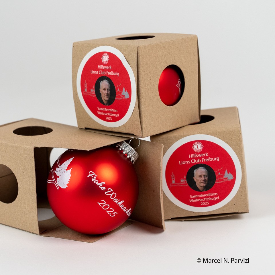 Drei rote Weihnachtskugeln mit weißen Motiven und der Aufschrift Frohe Weihnachten 2023 befinden sich in Pappschachteln, die jeweils mit einem Aufkleber mit dem Foto einer Person und den Aufschriften "Lions Club Freiburg" und "Sammelkollektion Weihnachtskugel 2023" versehen sind.