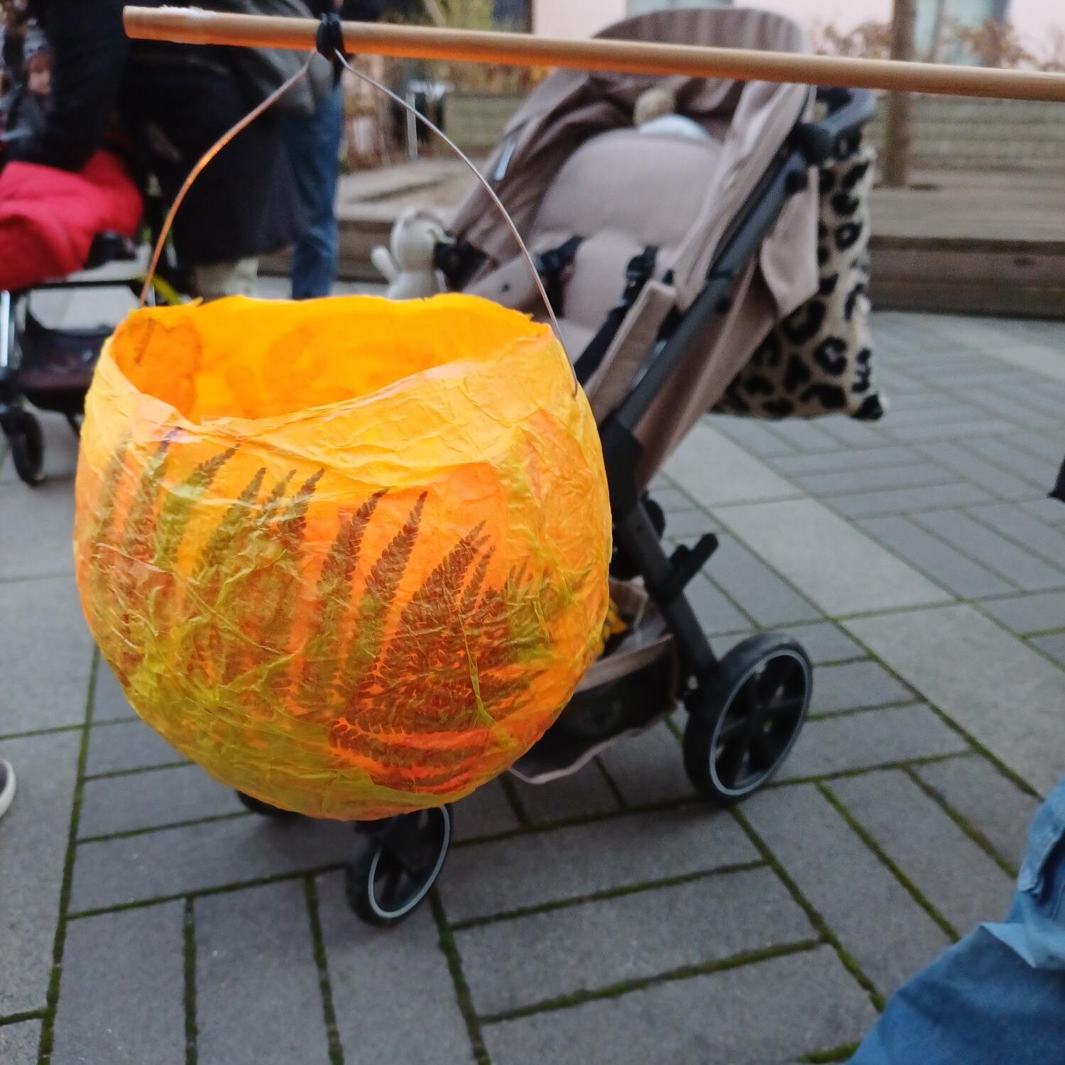 Eine leuchtend orangefarbene, mit Blattmustern verzierte Papierlaterne hängt an einem Stock, der an einem Kinderwagen befestigt ist. Eine Person in Jeans steht in der Nähe auf einem gepflasterten Gehweg.