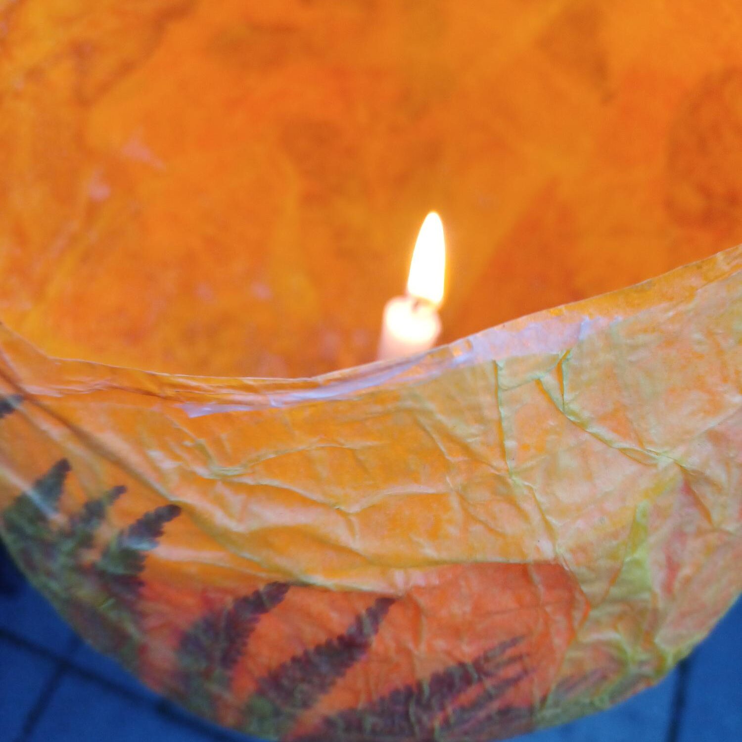 Eine brennende Kerze brennt in einer zerknitterten, orangefarbenen, durchscheinenden Papierlaterne, die mit Blattmustern verziert ist und auf einer Oberfläche aus quadratischen grauen Fliesen steht.