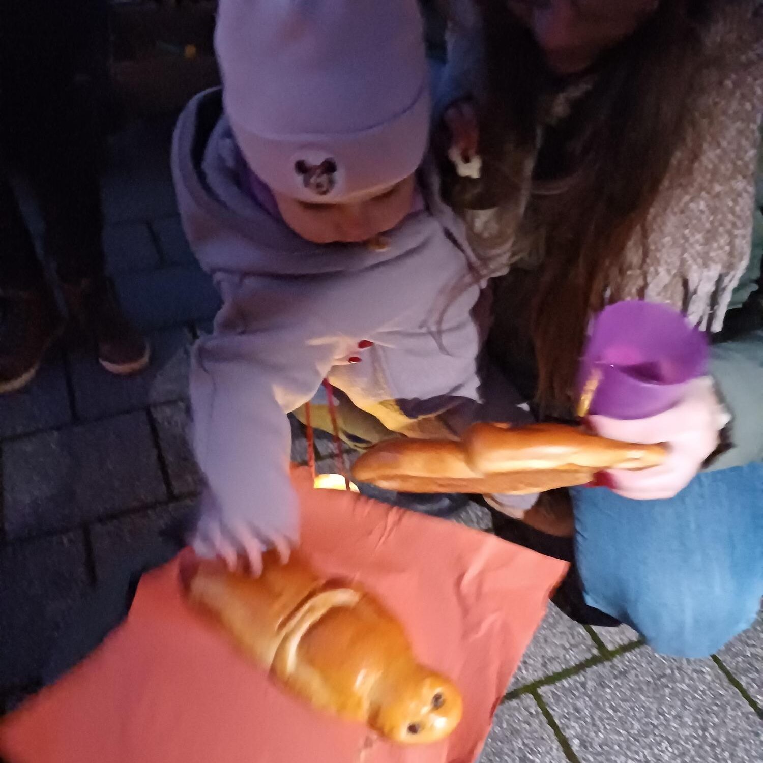 Ein kleines Kind mit einem rosa Hut greift nach einem Brot in Form einer Figur, die von einem Erwachsenen auf rotem Papier gehalten wird. Der Erwachsene hält eine andere Brotfigur und eine violette Laterne in der Hand. Sie befinden sich draußen auf einer gepflasterten Fläche.