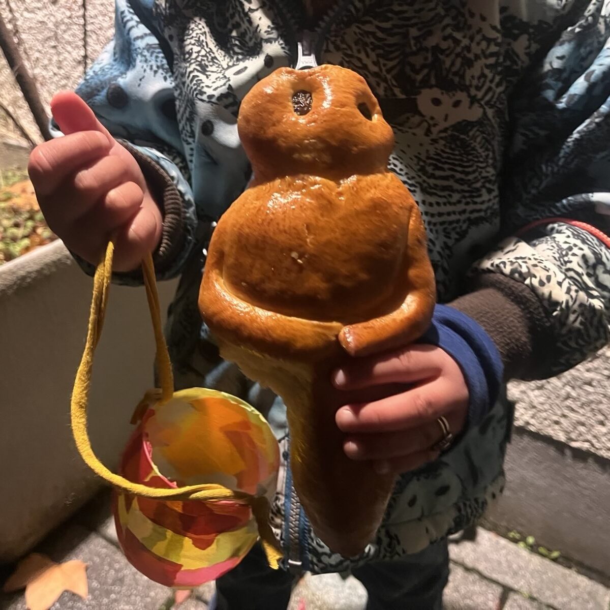 Ein Kind mit einem süßen Brot in Form eines Menschen in der einen Hand und einer bunten Laterne mit gelbem Henkel in der anderen, das in einer gemusterten Jacke im Freien steht.