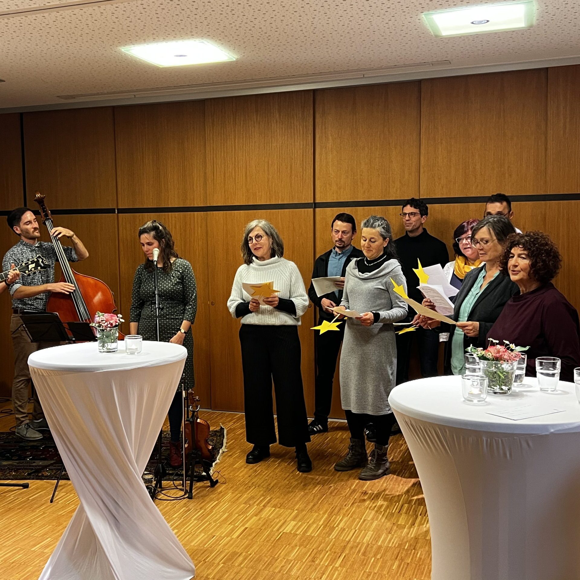 Eine Gruppe von Menschen steht in einem Raum mit Holzwänden, einige singen von Notenblättern, andere spielen Gitarre und Kontrabass. Im Vordergrund stehen zwei hohe Cocktailtische mit Getränken und Blumen.