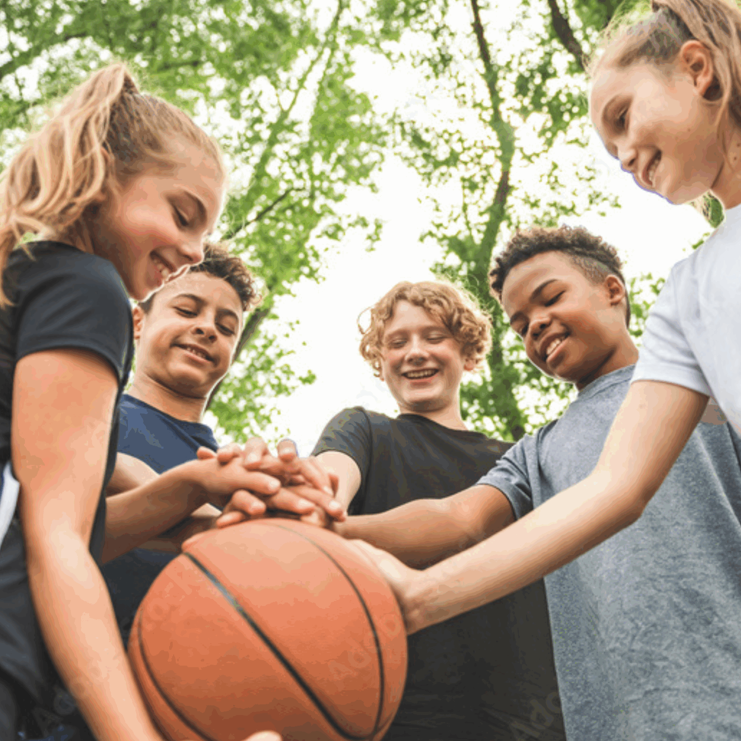 Fünf Kinder stehen im Freien in einem Kreis, lächeln und legen ihre Hände gemeinsam auf einen Basketball, zeigen Teamwork und Kameradschaft unter grünen Bäumen.
