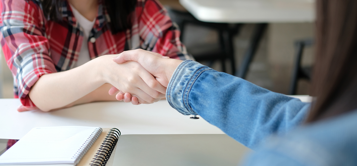 Zwei Personen schütteln sich über einen Tisch hinweg die Hände. Die eine trägt ein rot kariertes Hemd, die andere eine blaue Jeansjacke. Auf dem Tisch liegen ein Notizbuch und ein Stift, was auf ein Treffen oder ein Vorstellungsgespräch hindeutet.
