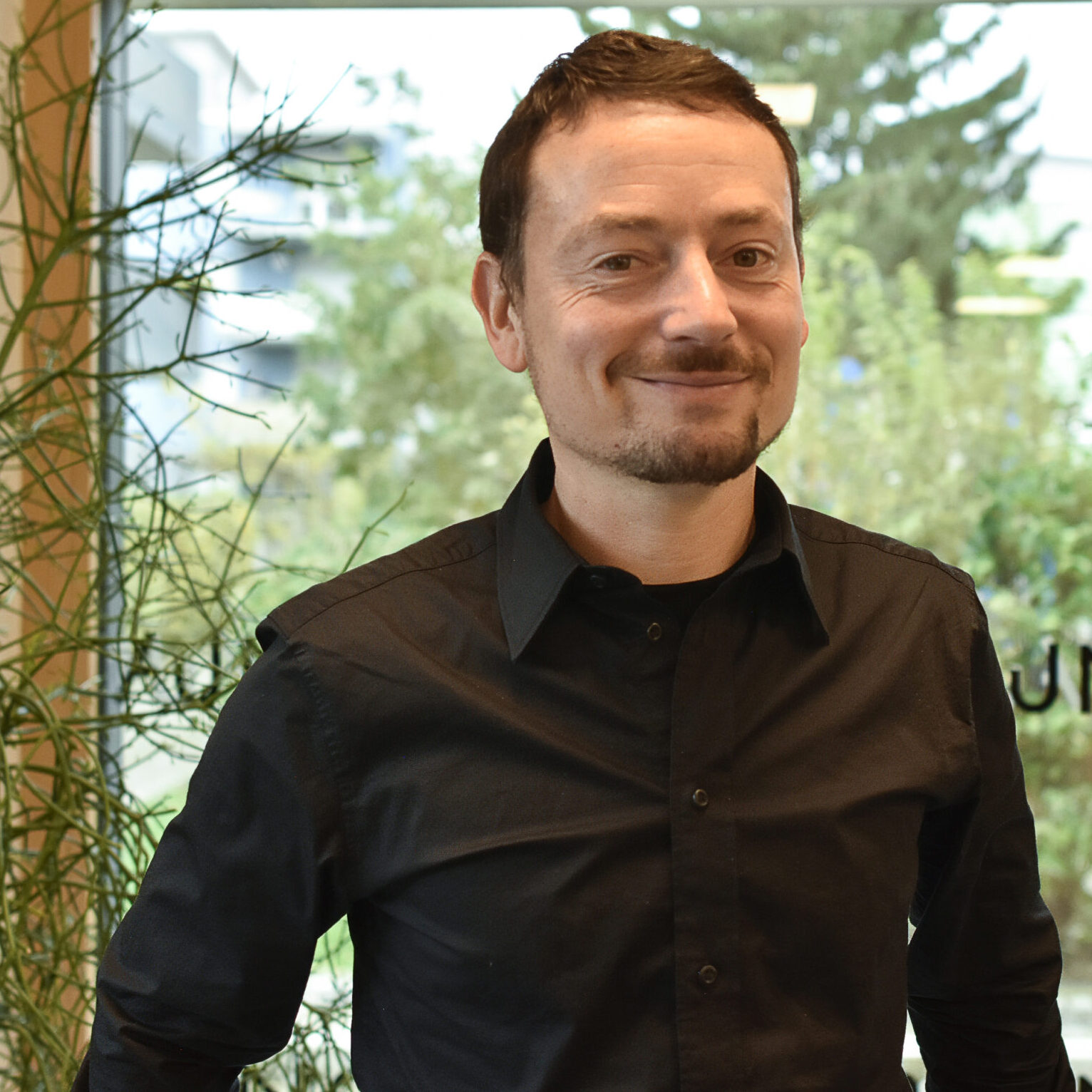 Ein Mann mit kurzen braunen Haaren und gestutztem Bart, der ein schwarzes Button-up-Hemd trägt, steht in einem Innenraum vor einem großen Fenster, aus dem Grünflächen zu sehen sind. Er lächelt sanft in die Kamera.
