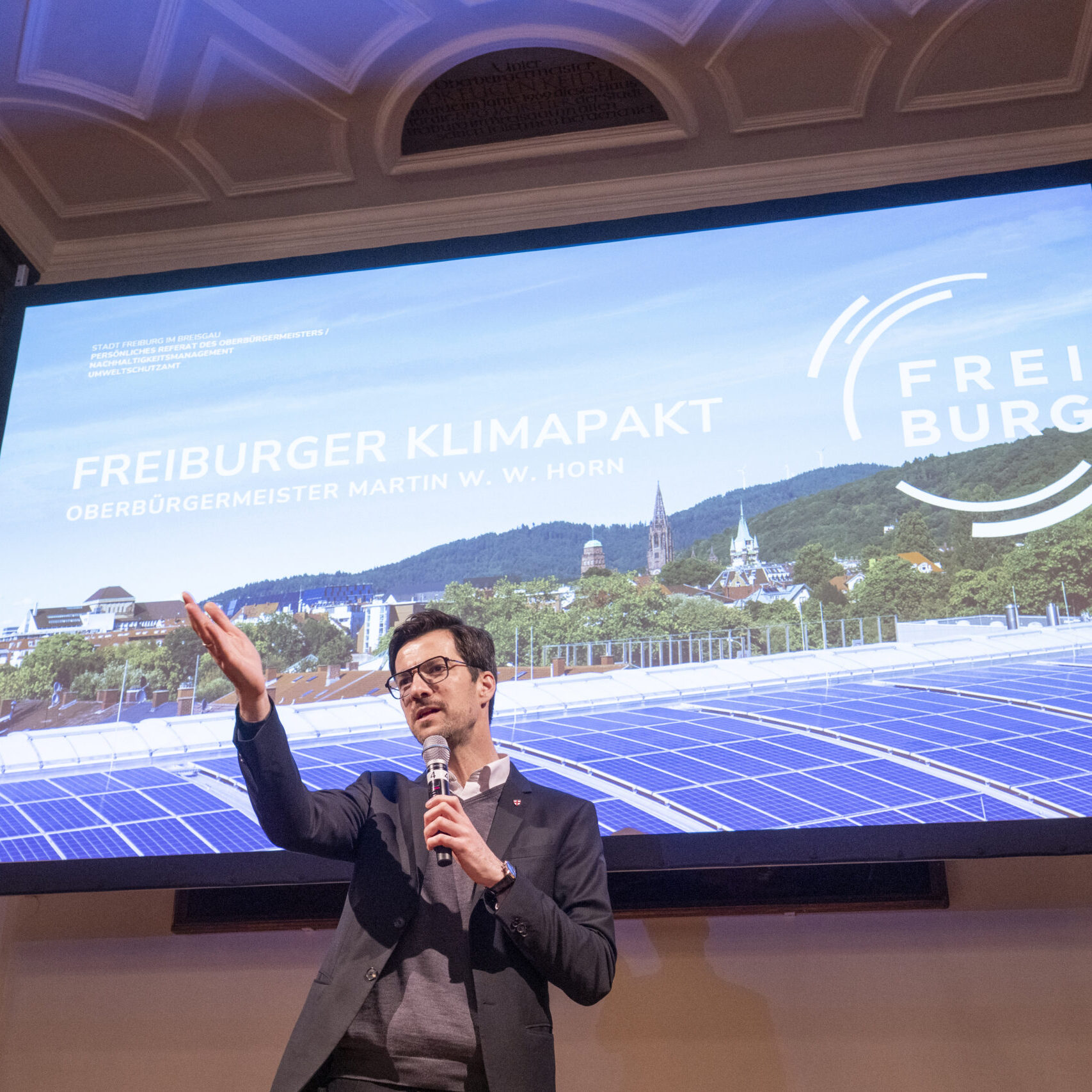 Ein Mann im Anzug spricht auf der Bühne mit einem Mikrofon und gestikuliert nach oben. Hinter ihm ist ein großer Bildschirm zu sehen, auf dem der Freiburger Klimapakt mit Bildern von Sonnenkollektoren und dem Freiburger Stadtbild zu sehen ist.