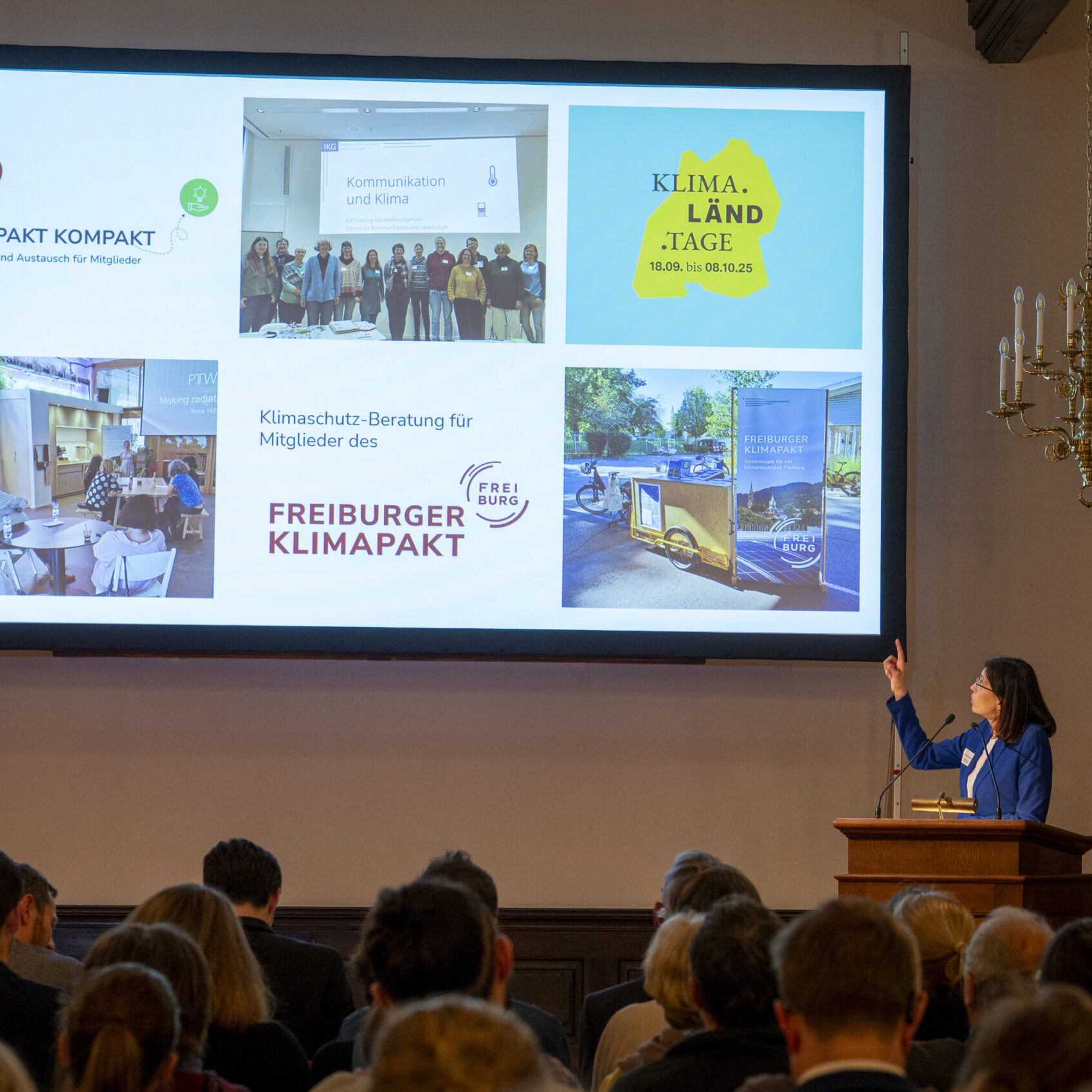 Eine Frau in einem blauen Anzug präsentiert dem Publikum in einem feierlichen Saal mit Kronleuchtern Dias über Klimainitiativen, während sie auf einen Bildschirm zeigt, auf dem verschiedene klimabezogene Bilder und Logos zu sehen sind.