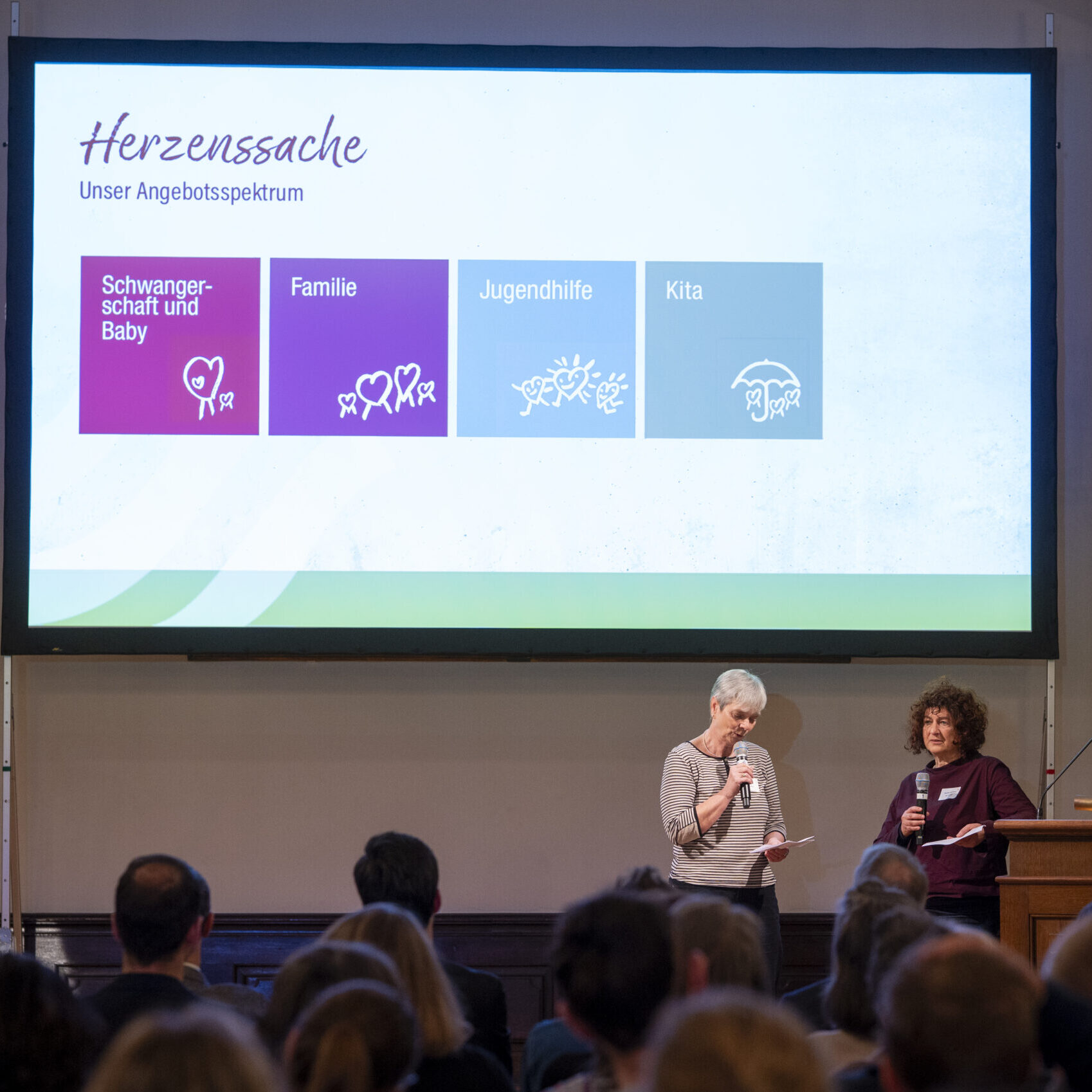 Zwei Frauen stehen auf der Bühne und sprechen zu den Zuhörern. Hinter ihnen ist eine große Präsentationsfolie zu sehen, auf der Symbole und deutscher Text über Dienstleistungen in den Bereichen Schwangerschaft, Familie, Jugendhilfe und Tagesbetreuung zu sehen sind. Weibliche Inspiration. Stark für Frauen. Stark für Menschen.