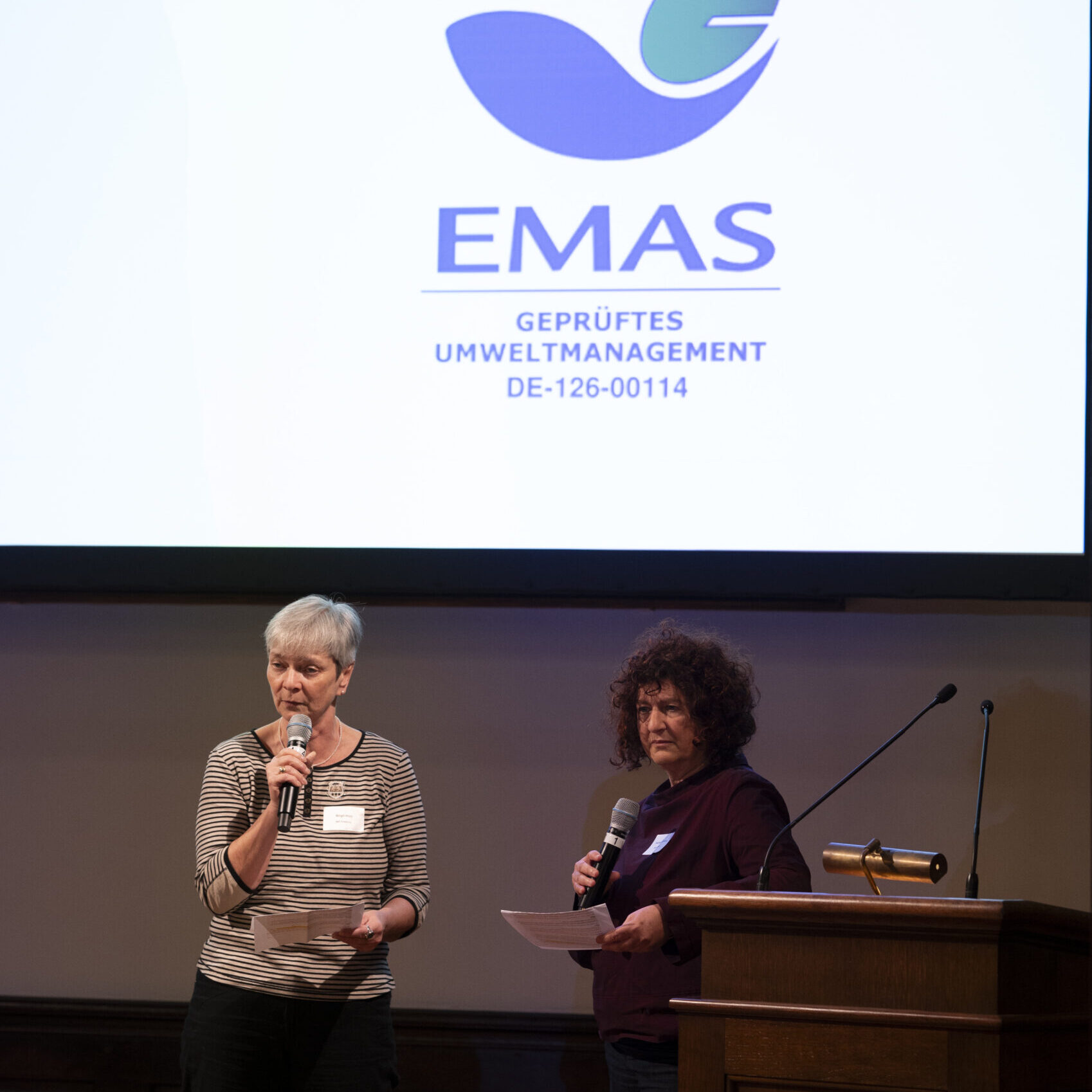 Zwei Frauen stehen auf der Bühne und sprechen in Mikrofone vor einem großen Bildschirm mit dem EMAS-Logo und dem Text EMAS GEPRÜFTES UMWELTMANAGEMENT DE-126-00114.