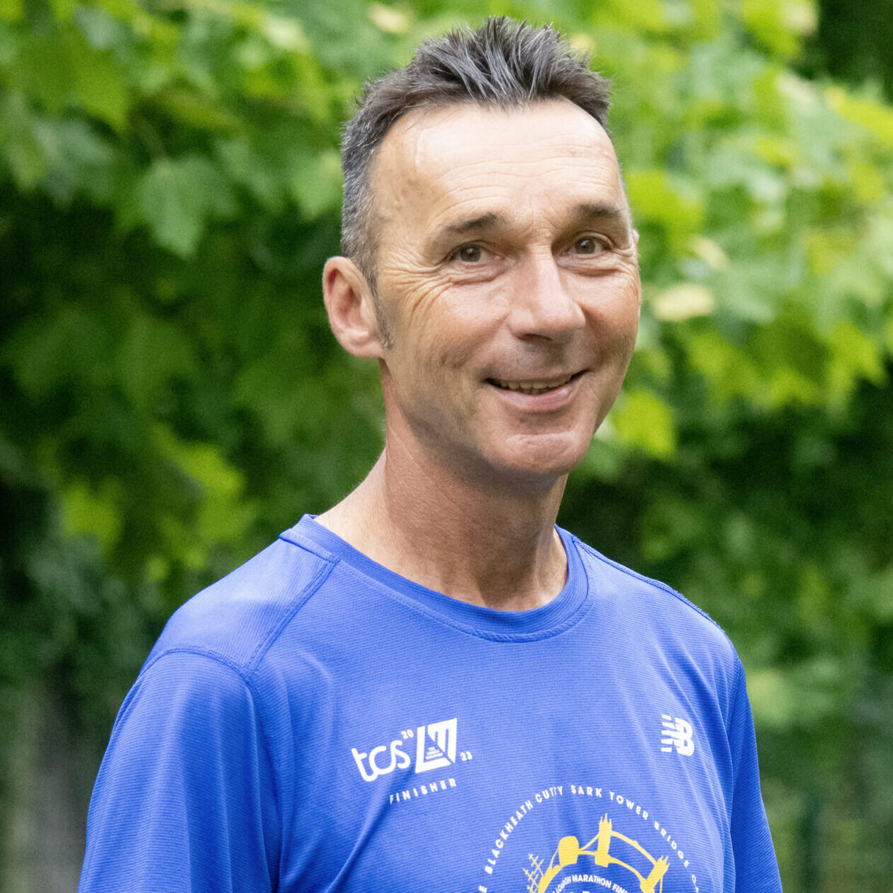 Ein Mann in einem blauen Sporthemd steht im Freien in einem üppigen, grünen Park und lächelt in die Kamera. Bäume und Laub füllen den Hintergrund.