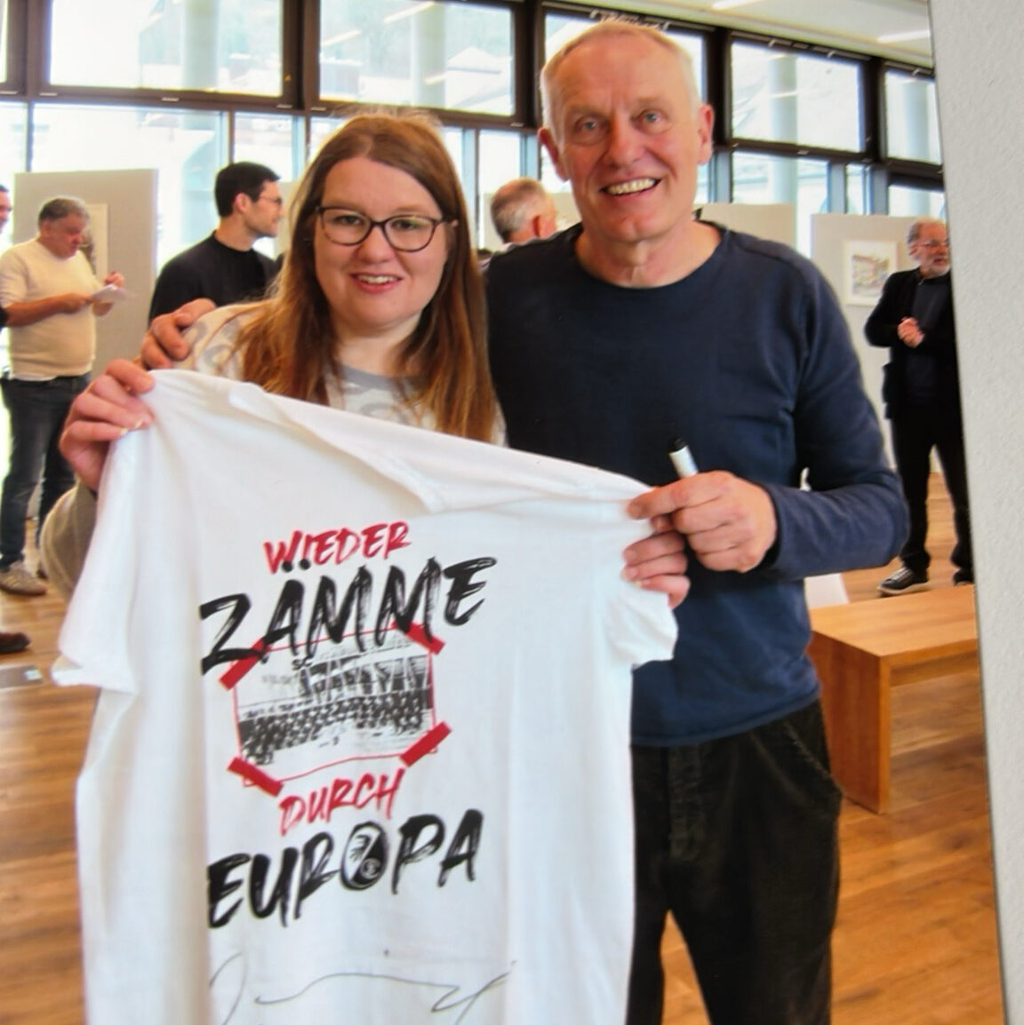 Zwei Personen lächeln in einem Haus und halten ein weißes T-Shirt, auf dem in fetten Buchstaben "Wieder Zamme Durch Europa" steht. Im Hintergrund sind andere Menschen und große Fenster zu sehen.
