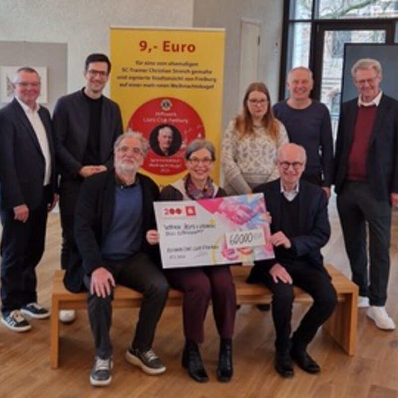 Eine Gruppe von neun Personen posiert in einem Innenraum, einige stehend, andere sitzend, und hält einen großen Scheck über 60.000 Euro in der Hand. Hinter ihnen ist ein gelbes und rotes Plakat zu sehen und an den Wänden hängen Kunstwerke im Stil einer Galerie.