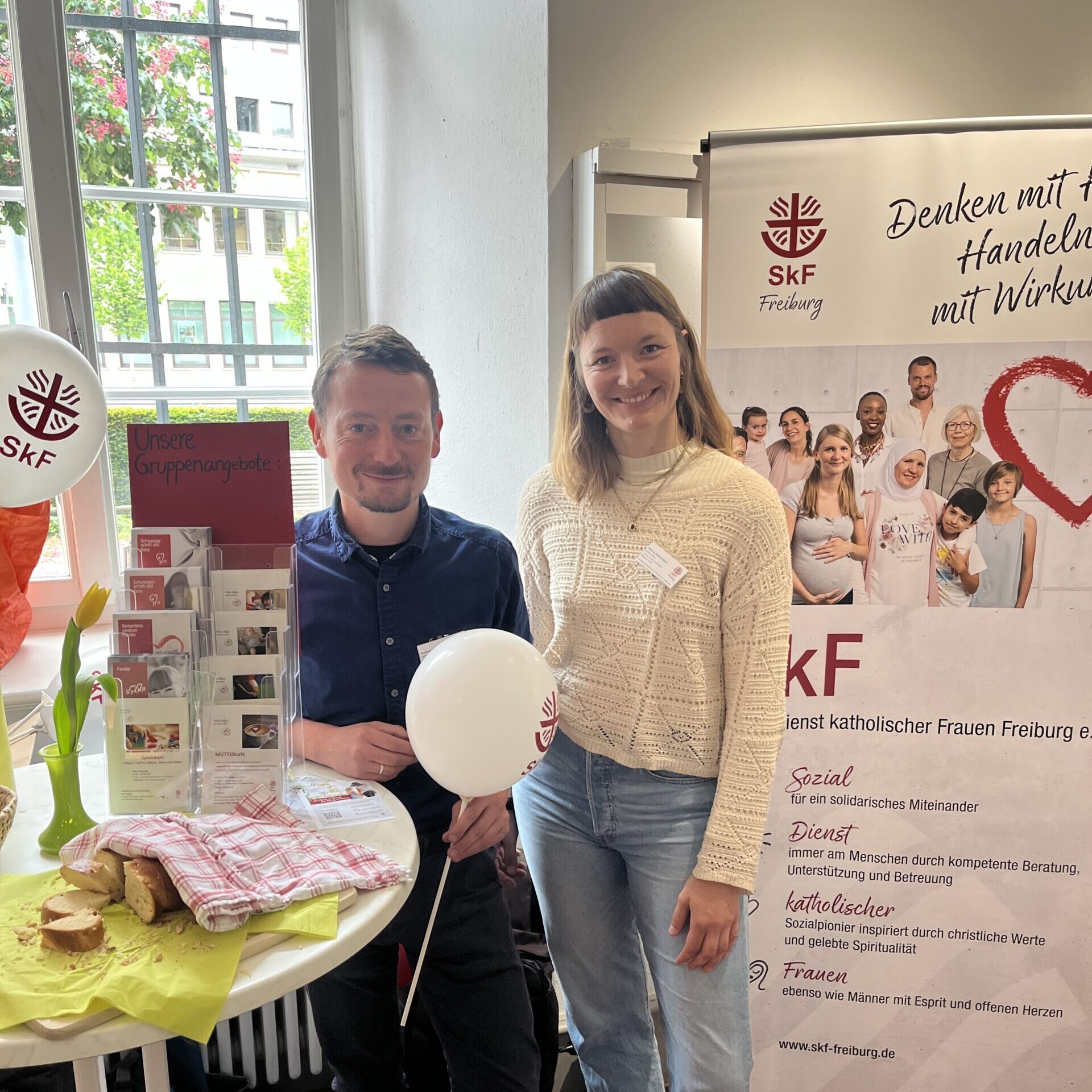 Zwei Personen stehen lächelnd an einem Stand mit Infomaterial, Luftballons und Snacks. Hinter ihnen hängt ein Banner des SkF Freiburg mit Bildern von Frauen und Texten über soziale Dienste. Weibliche Inspiration. Stark für Frauen. Stark für Menschen.