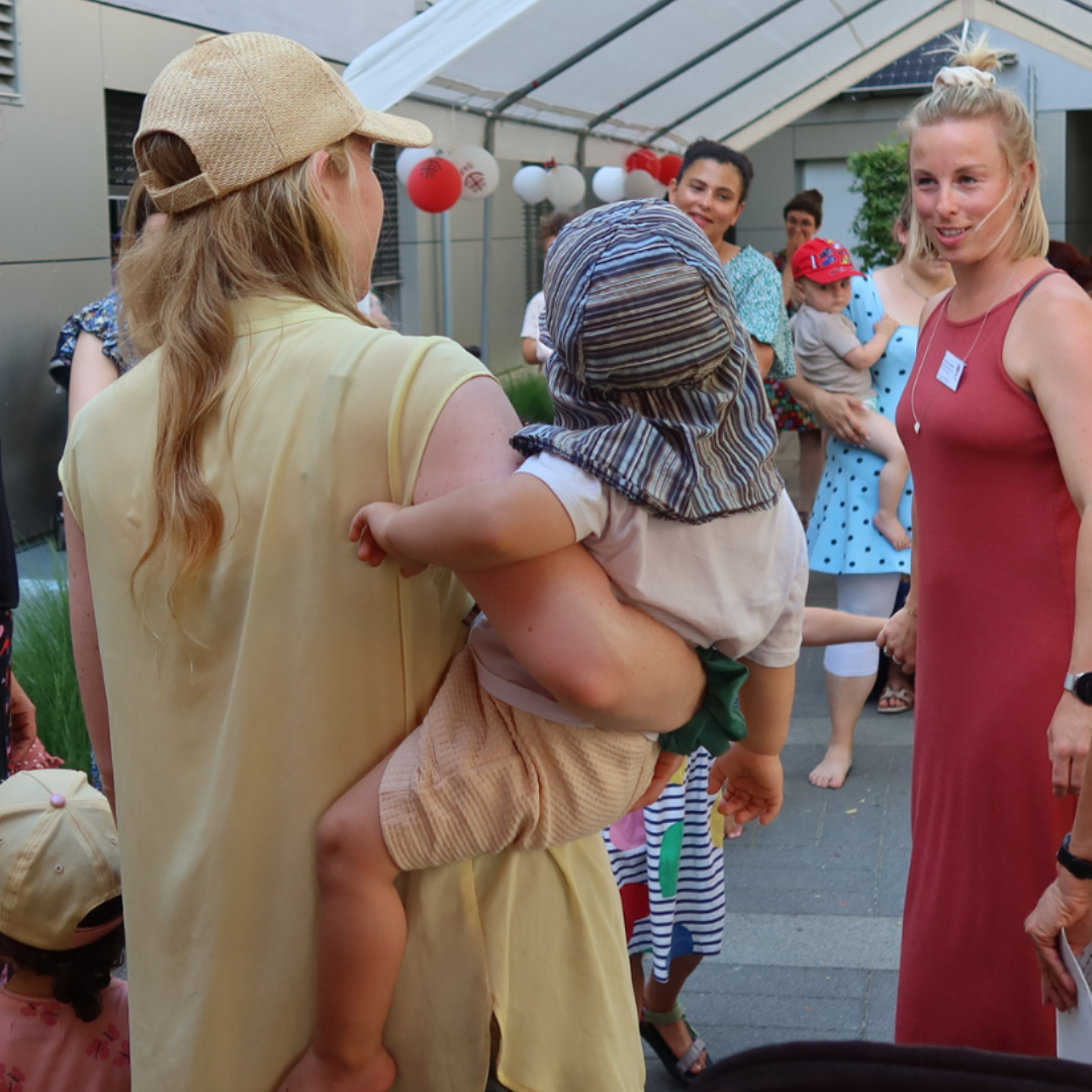 Eine Gruppe von Erwachsenen und Kindern versammelt sich im Freien unter einem mit roten und weißen Laternen geschmückten Vordach. Eine Frau hält ein Kind mit einer gestreiften Mütze im Arm, während eine andere Frau in einem roten Kleid daneben lächelt.