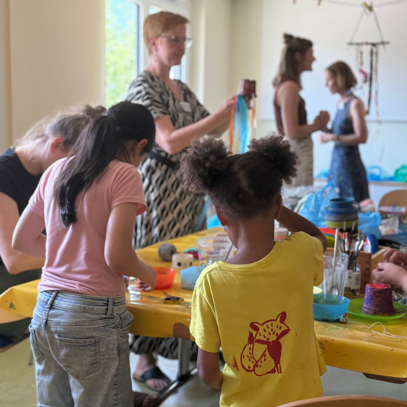 Mehrere Kinder und Erwachsene haben sich um einen gelben Tisch versammelt und basteln. Im Hintergrund stehen zwei Frauen und unterhalten sich, während andere sitzen oder an ihren Projekten arbeiten. Der Raum ist von Tageslicht gut beleuchtet.