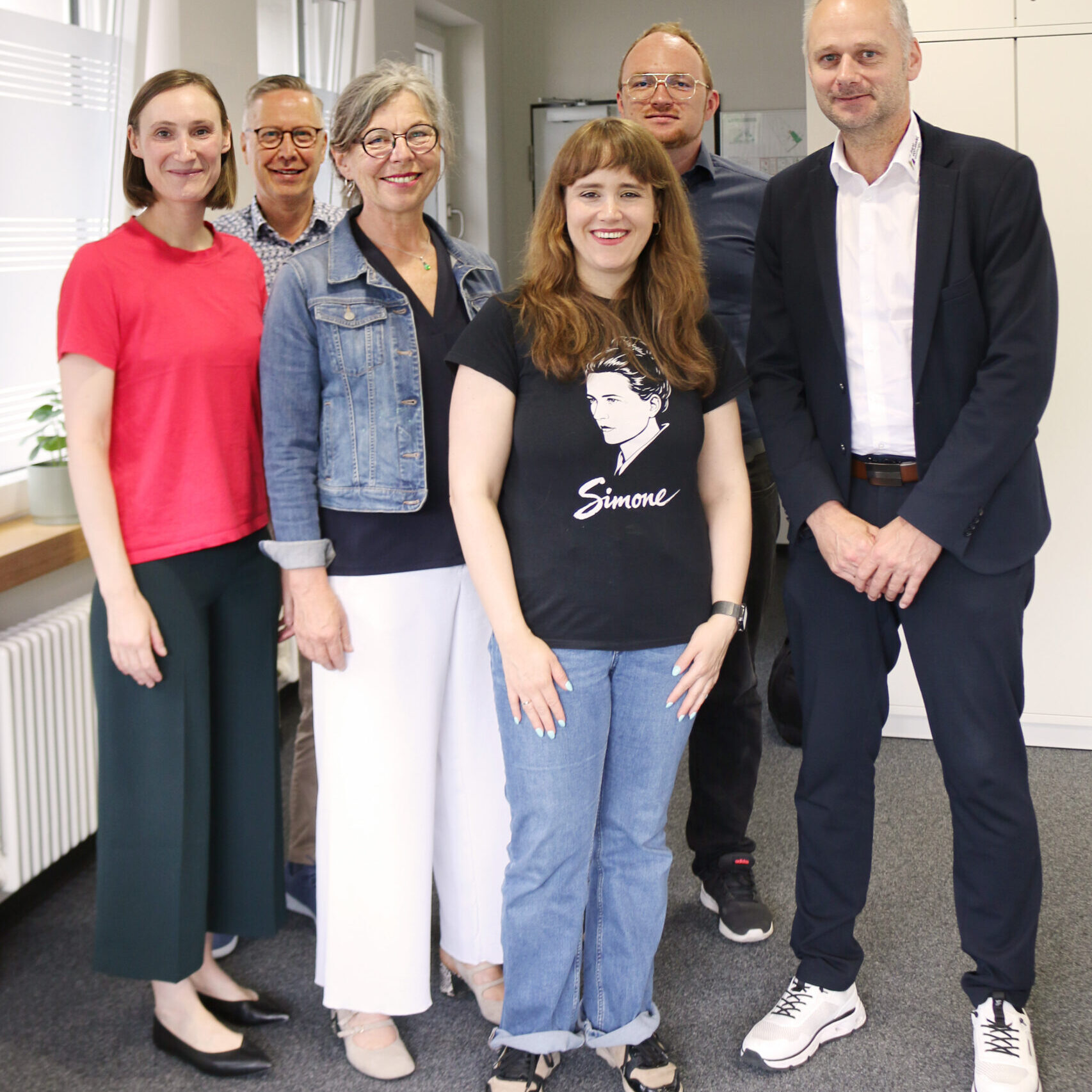 Sechs Personen stehen lächelnd in einem Büro, teilweise in Freizeitkleidung, teilweise in Geschäftskleidung. Eine Person in der Mitte trägt ein schwarzes T-Shirt mit einem Porträt und dem Namen „Simone“ darauf. Die Gruppe wirkt freundlich und entspannt. SkF Spenden-Projekte