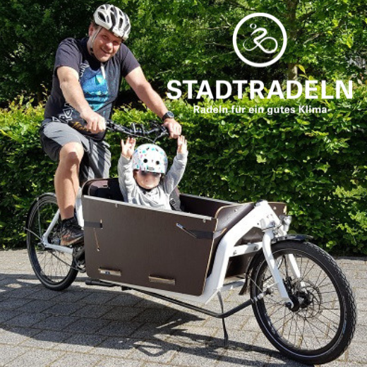 Ein Mann mit Helm fährt auf einem Lastenrad, auf dessen vorderem Korb ein Kind sitzt; beide lächeln. Der Text lautet „STADTRADELN – Radeln für ein gutes Klima“, darüber ist ein Fahrrad-Logo zu sehen. Im Hintergrund ist üppiges Grün zu sehen. Weibliche Inspiration. Stark für Frauen. Stark für Menschen.