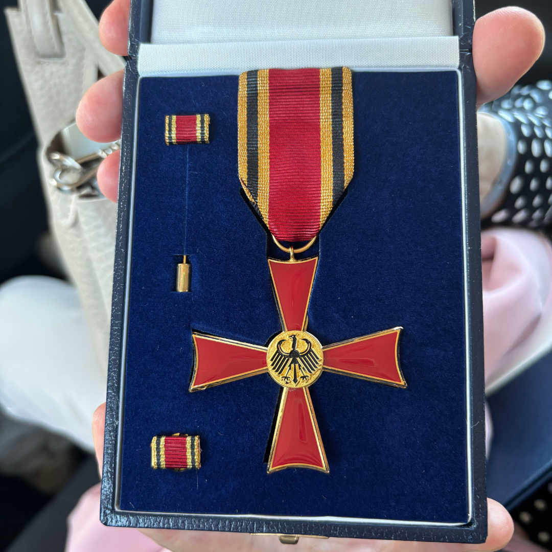 Eine Hand hält eine blaue Schachtel, in der sich eine rot-goldene, kreuzförmige Medaille mit einem gestreiften Band sowie zwei dazu passende Bandstreifen befinden.