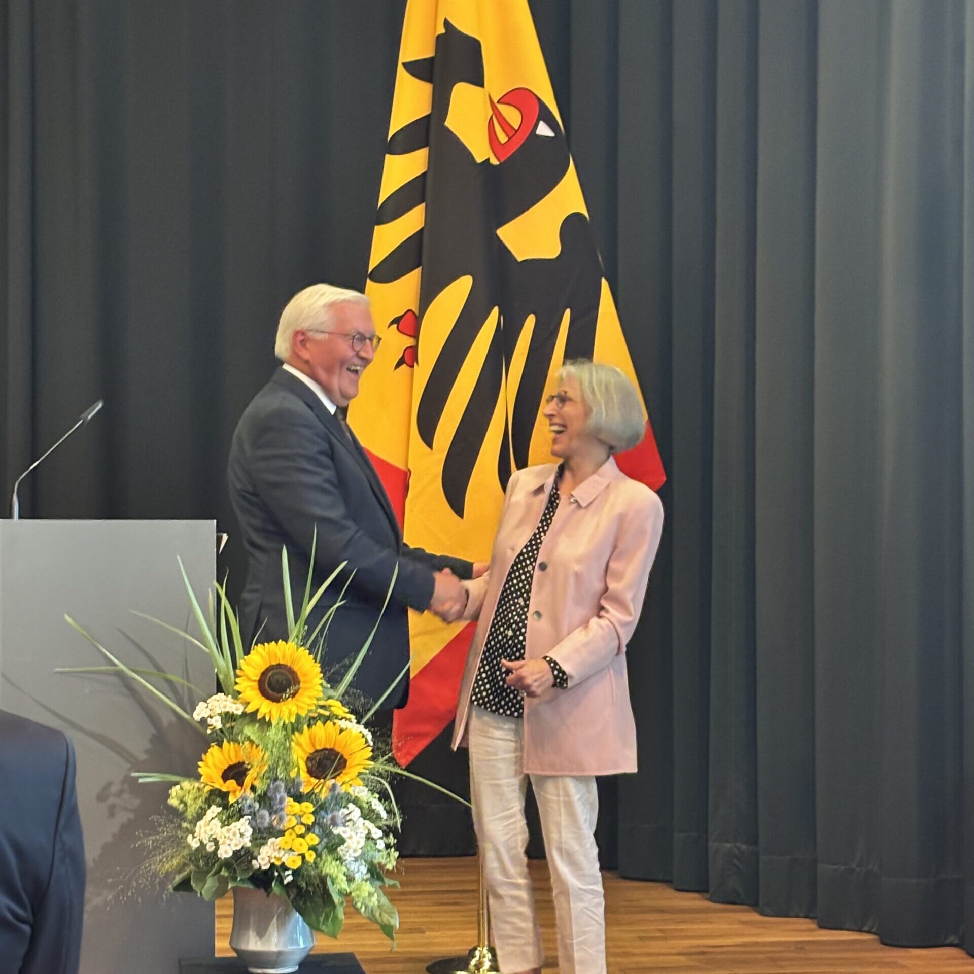 Ein Mann und eine Frau geben sich auf einer Bühne, auf der eine große deutsche Flagge und ein mit Sonnenblumen geschmücktes Podium zu sehen sind, die Hand und lächeln. Im Hintergrund sind schwarze Vorhänge zu sehen.