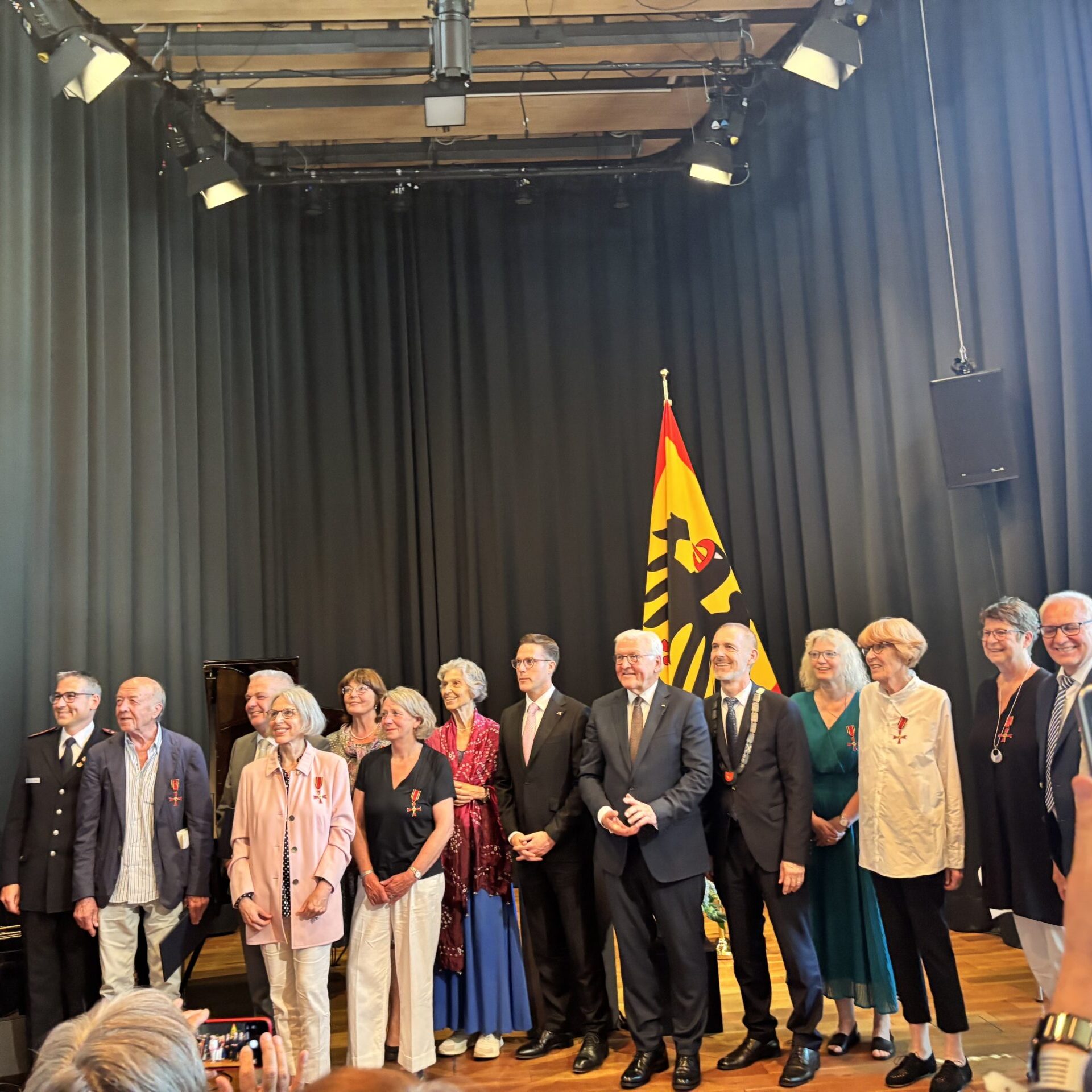 Eine Gruppe von Menschen posiert gemeinsam auf einer Bühne vor einer Flagge, gekleidet in formelle und halbformelle Kleidung, im Schein der Bühnenbeleuchtung, während jemand im Vordergrund mit einem Handy ein Foto macht.