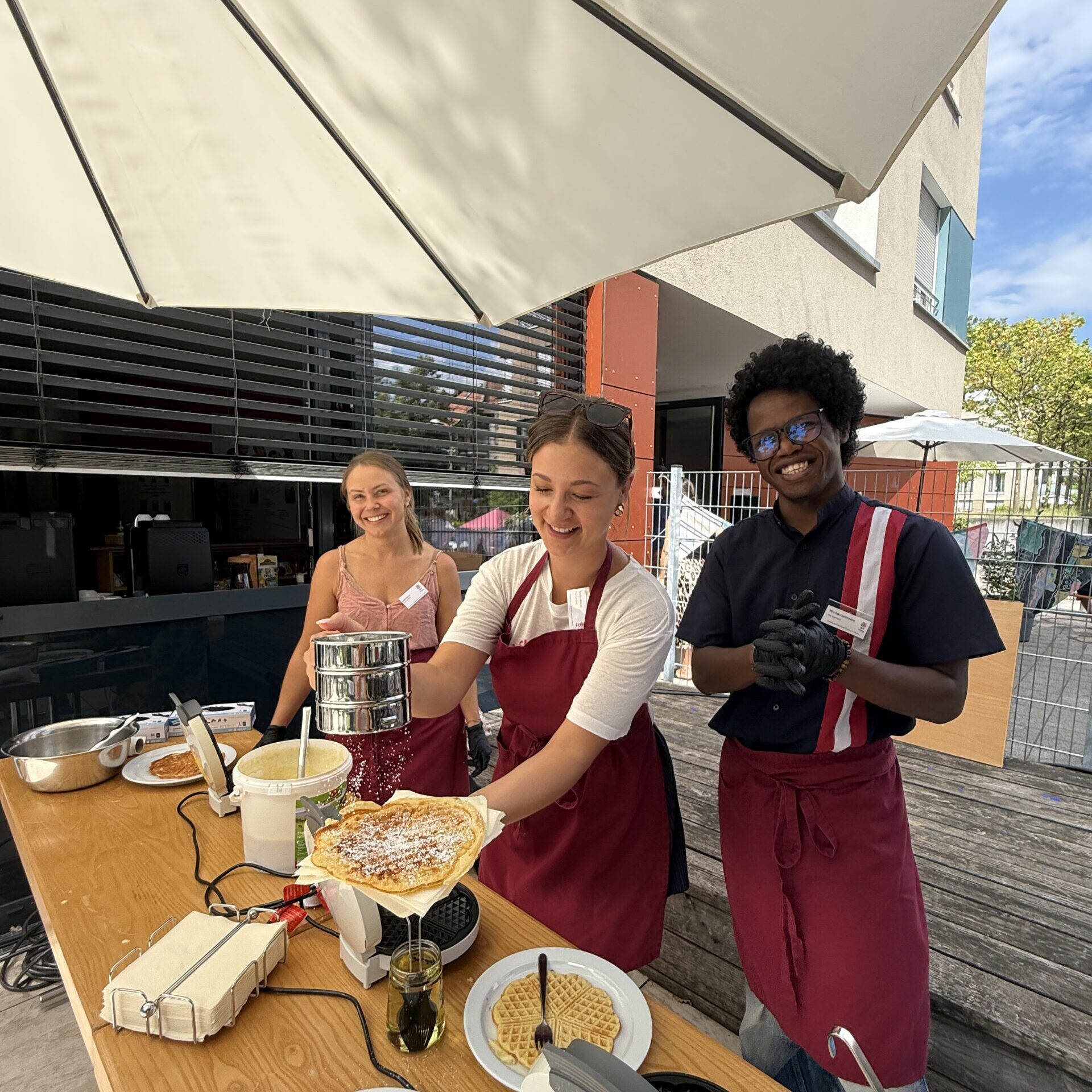 Drei Personen in Schürzen stehen an einem Tisch im Freien und backen Waffeln. Eine Frau lächelt, während sie eine Waffel in der Hand hält, eine weitere steht neben ihr, und ein Mann mit Handschuhen lächelt in die Kamera. Auf dem Tisch stehen Teller und Waffeleisen.