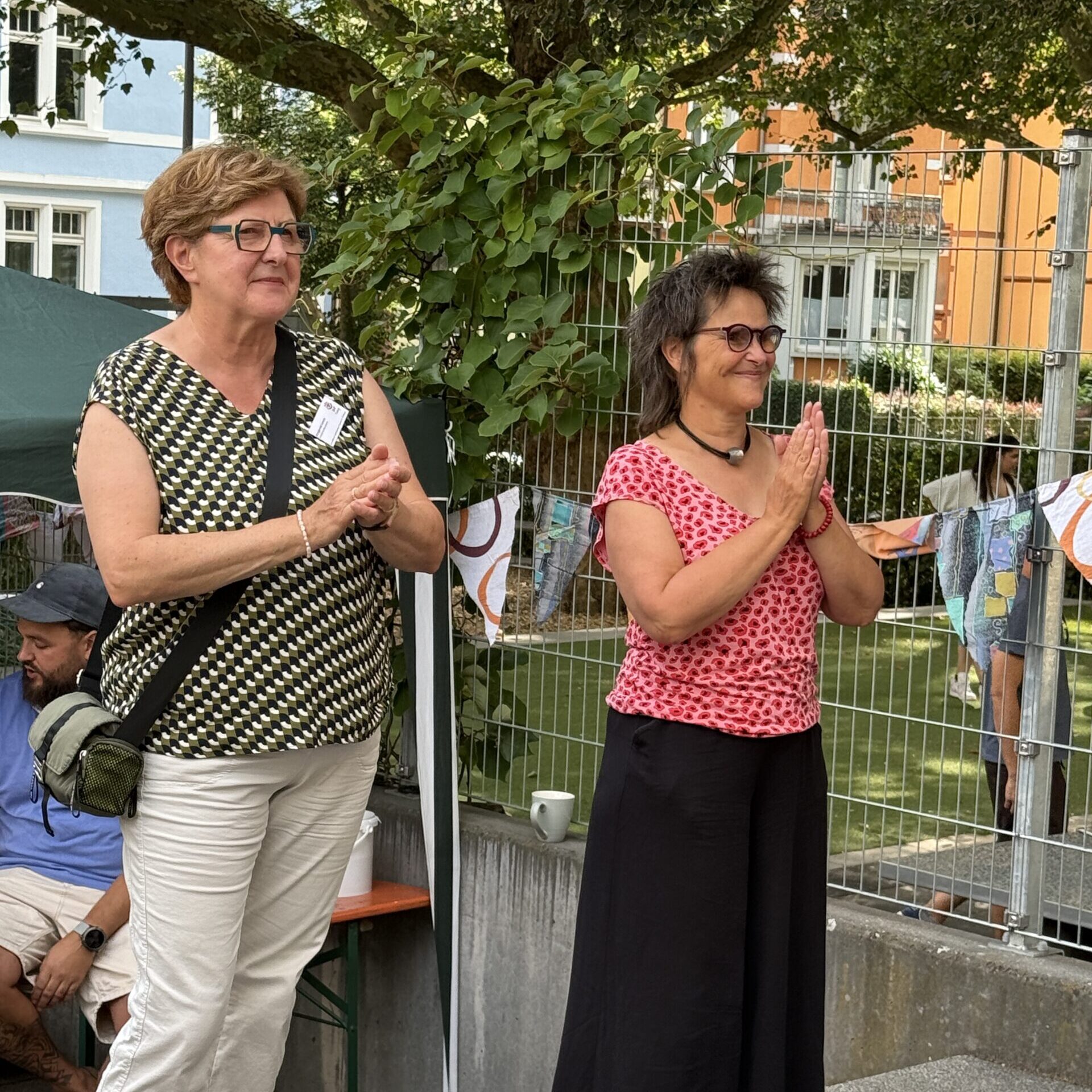 Zwei Frauen stehen im Freien in der Nähe eines Zauns und von Grünpflanzen, klatschen und lächeln. Die eine trägt eine Brille, ein gemustertes Oberteil und eine helle Hose; die andere hat ein rosa Oberteil und einen schwarzen Rock an. Im Hintergrund sind Menschen und festliche Dekorationen zu sehen.