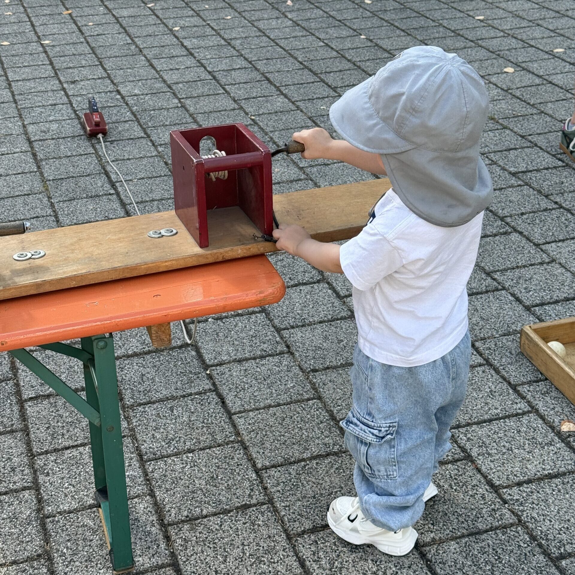 Ein kleines Kind, das einen Sonnenhut und Jeans trägt, dreht draußen an einer Kurbel eines roten Holzgeräts und steht dabei auf einem gepflasterten Untergrund in der Nähe von Laub.