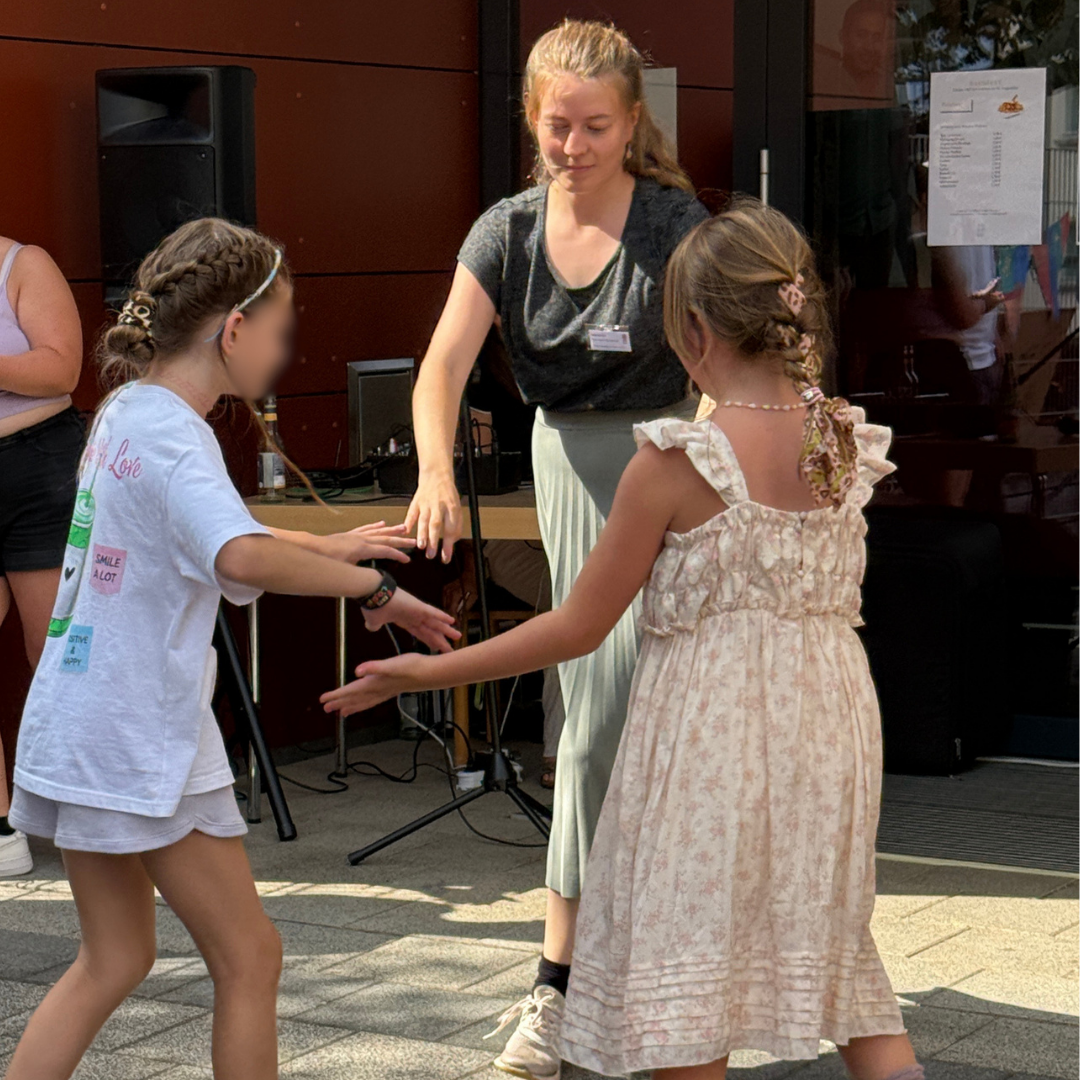 Eine Frau lächelt, während sie draußen mit zwei kleinen Mädchen steht, die spielen und Händchen halten; alle wirken glücklich und sind ganz in ihre Aktivität im Freien vertieft.