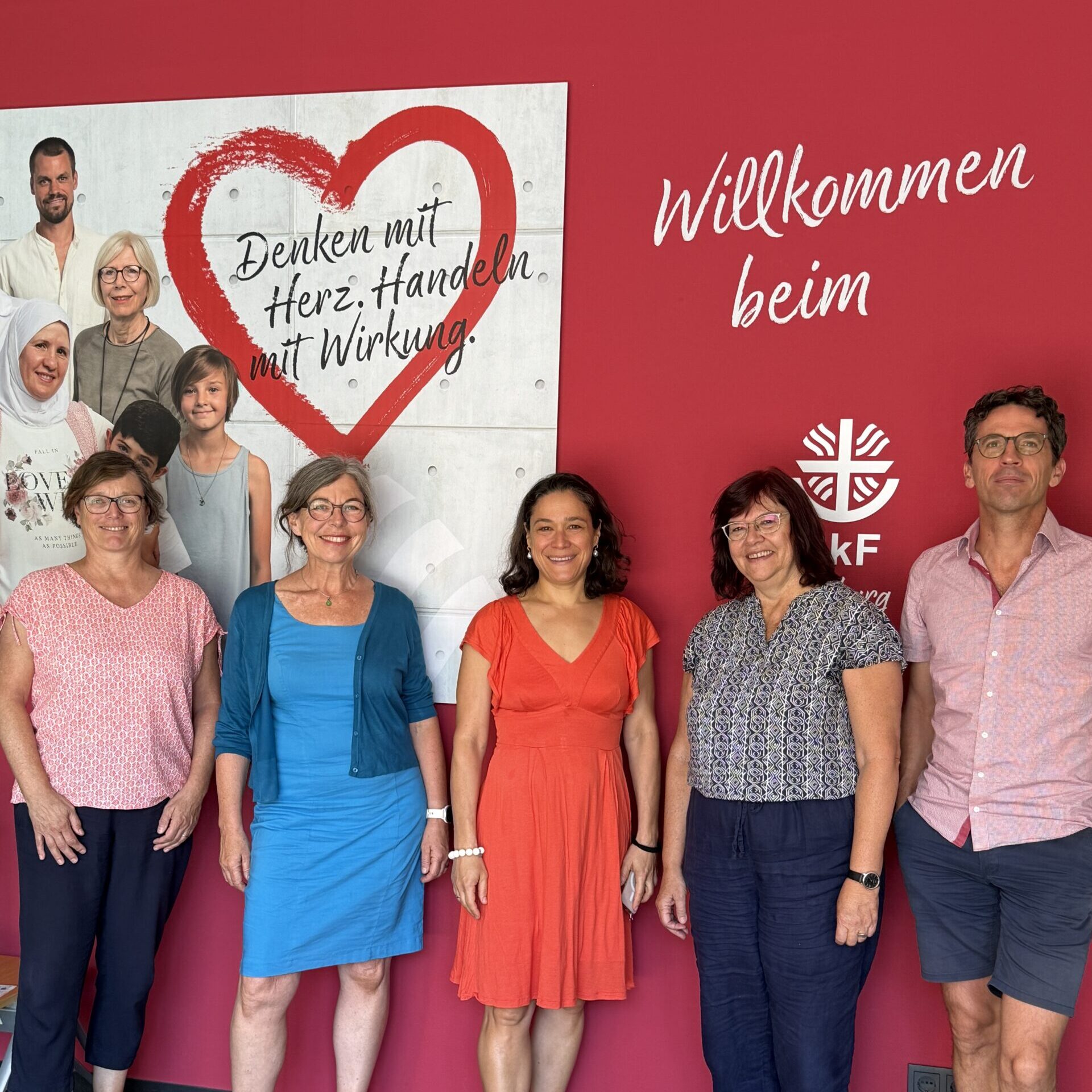 Fünf Erwachsene stehen lächelnd vor einer roten Wand, auf der ein Begrüßungstext und ein Plakat mit einem Herz sowie Gruppenfotos zu sehen sind. Vier Frauen und ein Mann posieren gemeinsam, alle in Sommerkleidung.