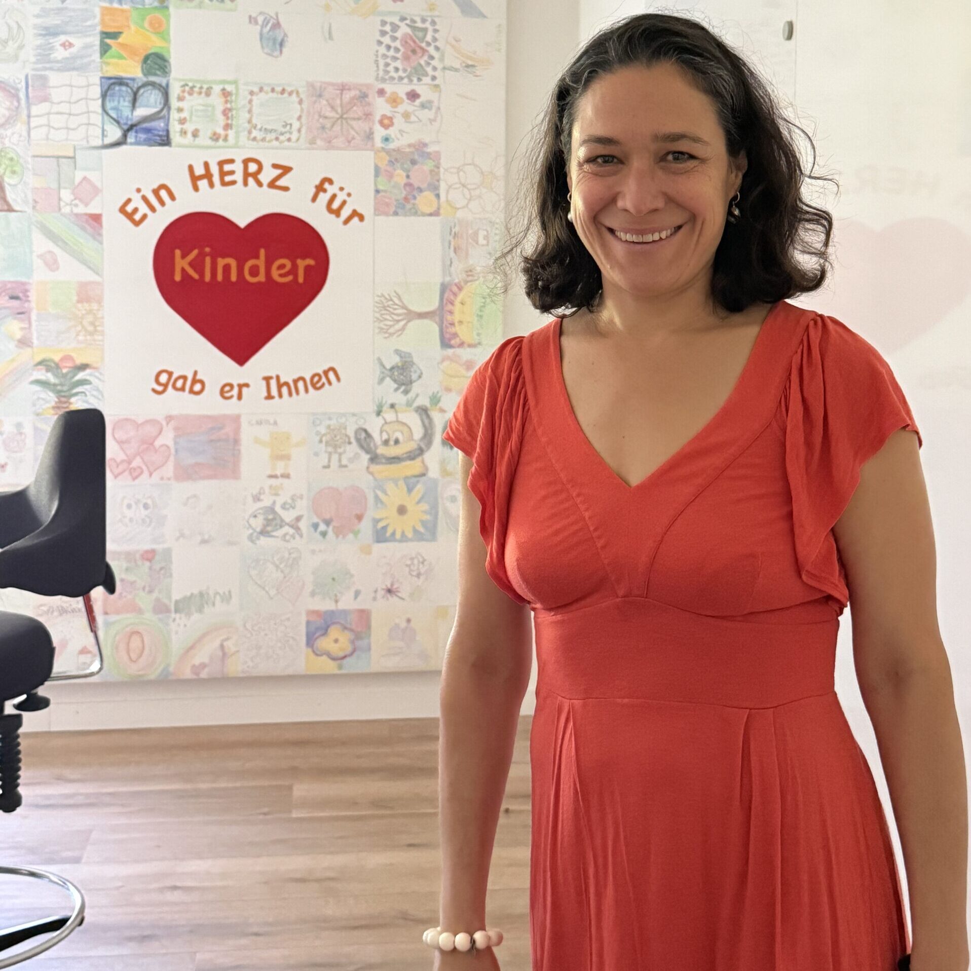 Eine Frau in einem roten Kleid lächelt in einem hellen Raum. Hinter ihr befindet sich eine bunte Wand mit Kinderzeichnungen und einem Schild, auf dem steht: „Ein HERZ für Kinder gab er Ihnen…“. Weibliche Inspiration. Stark für Frauen. Stark für Menschen.