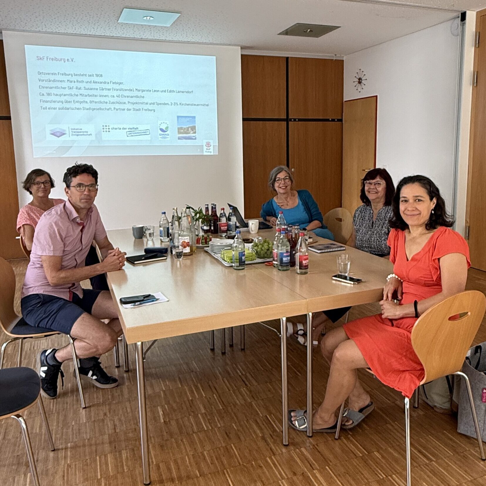 Sechs Personen sitzen mit Getränken und Unterlagen um einen Konferenztisch herum. Hinter ihnen wird eine Präsentation an die Wand projiziert. Der Raum hat Holzböden und geschlossene Holztüren.