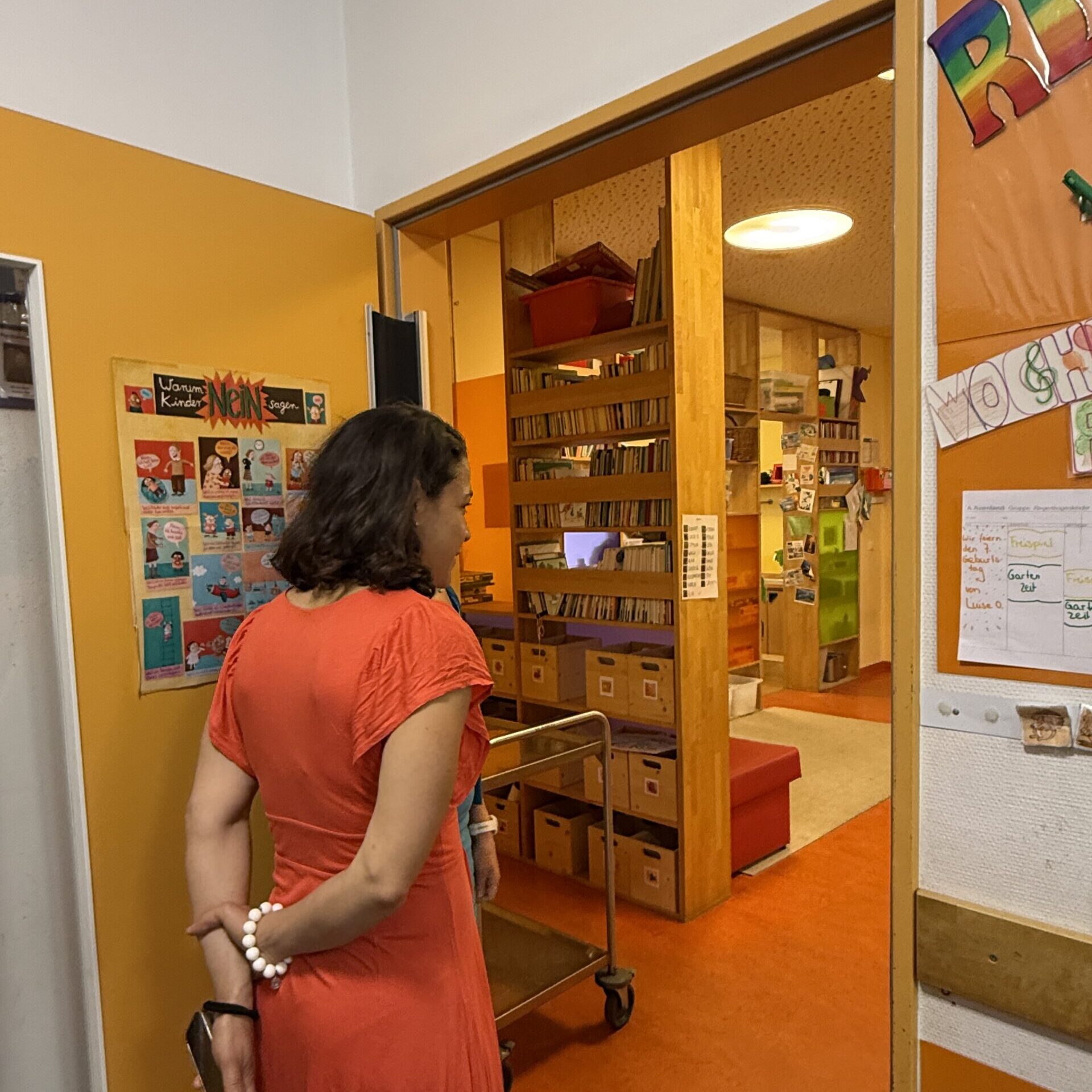 Eine Frau in einem orangefarbenen Kleid steht in der Tür eines hell erleuchteten Raums, der mit Bücherregalen und Kisten gefüllt ist; an den Wänden um sie herum hängen bunte Plakate und Zettel.