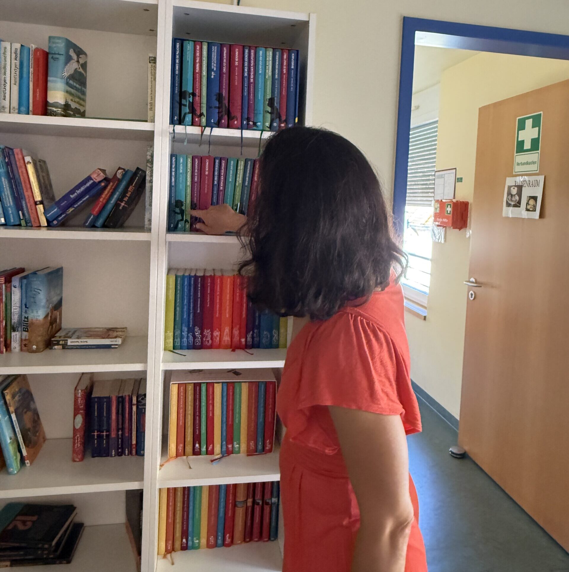 Eine Frau in einem roten Kleid steht neben einem Bücherregal und greift nach einem Buch mit bunten Buchrücken. Der Raum hat gelbe Wände, einen blauen Türrahmen und im Hintergrund ist an der Tür ein Erste-Hilfe-Zeichen zu sehen.