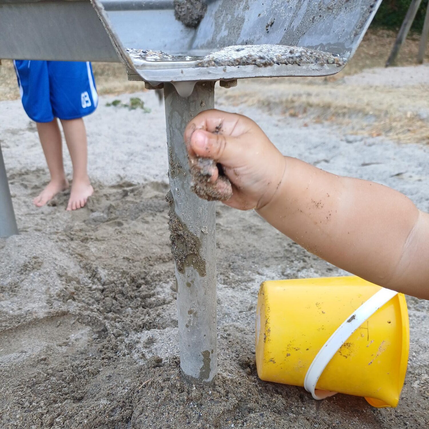 Auf einem Spielplatz greift die sandbedeckte Hand eines Kindes nach einer Metallstange. Ein gelber Eimer liegt daneben im Sand. Im Hintergrund steht ein weiteres barfüßiges Kind in blauen Shorts.