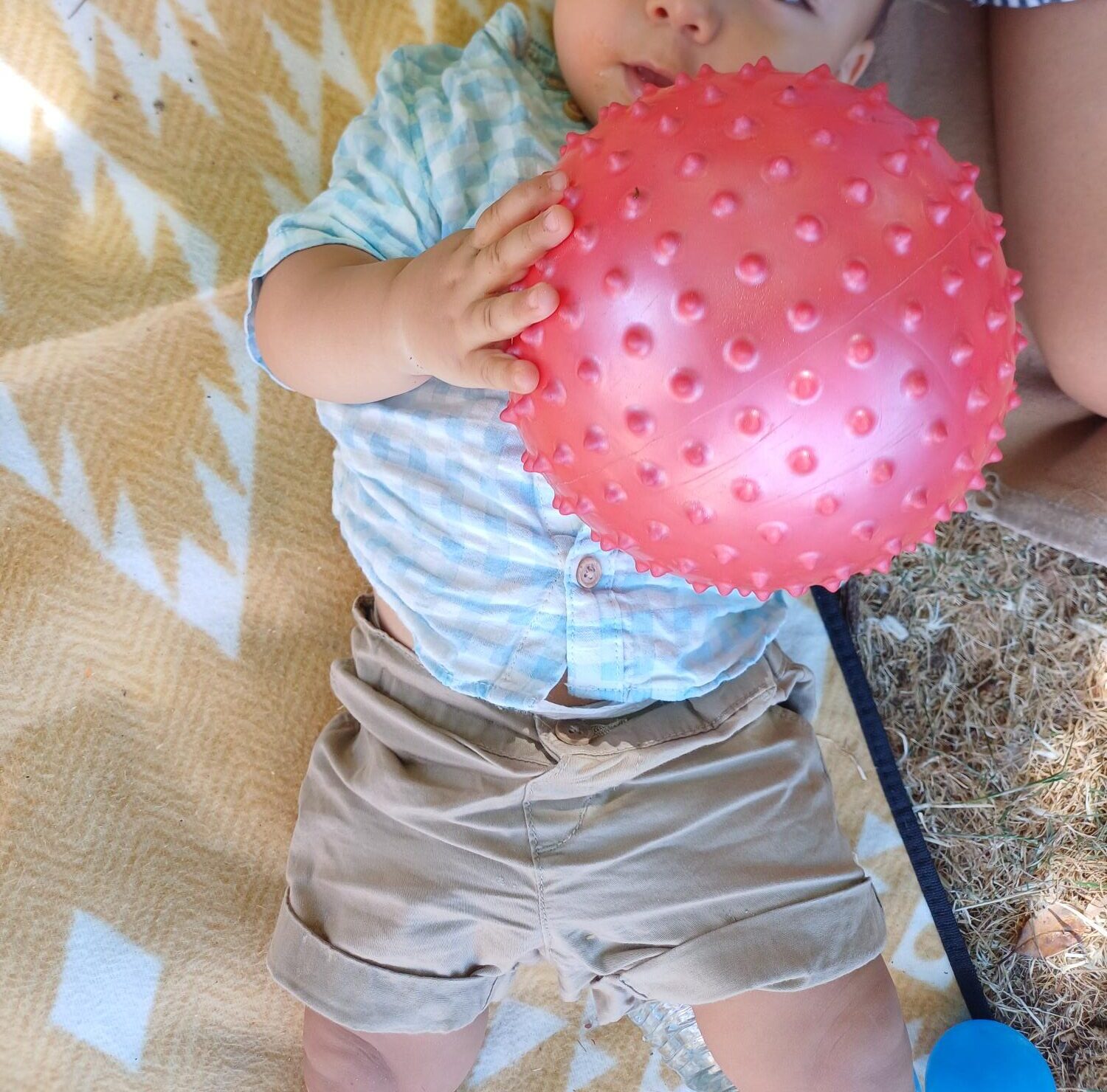Ein Baby liegt draußen auf einer gemusterten Decke und hält einen großen, rosa Ball mit Struktur in den Händen. Neben dem Baby ist der Arm eines Erwachsenen zu sehen.