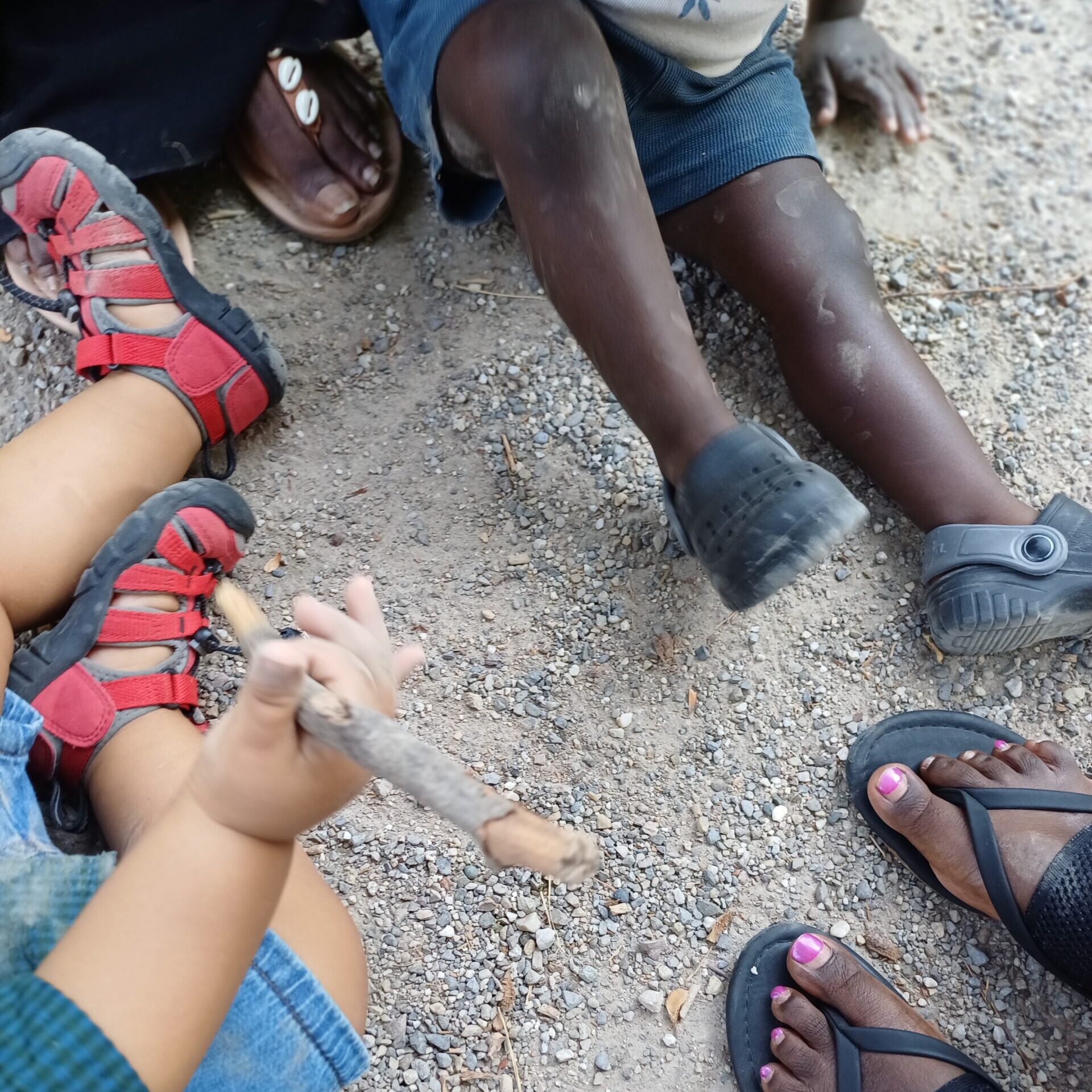 Kinder sitzen auf Kies, eines hält einen Stock in der Hand; sie tragen Sandalen und Shorts. Im Bild sind die Füße eines Erwachsenen mit lackierten Zehennägeln zu sehen, im Hintergrund ist eine weitere Person mit Schuhen zu erkennen.